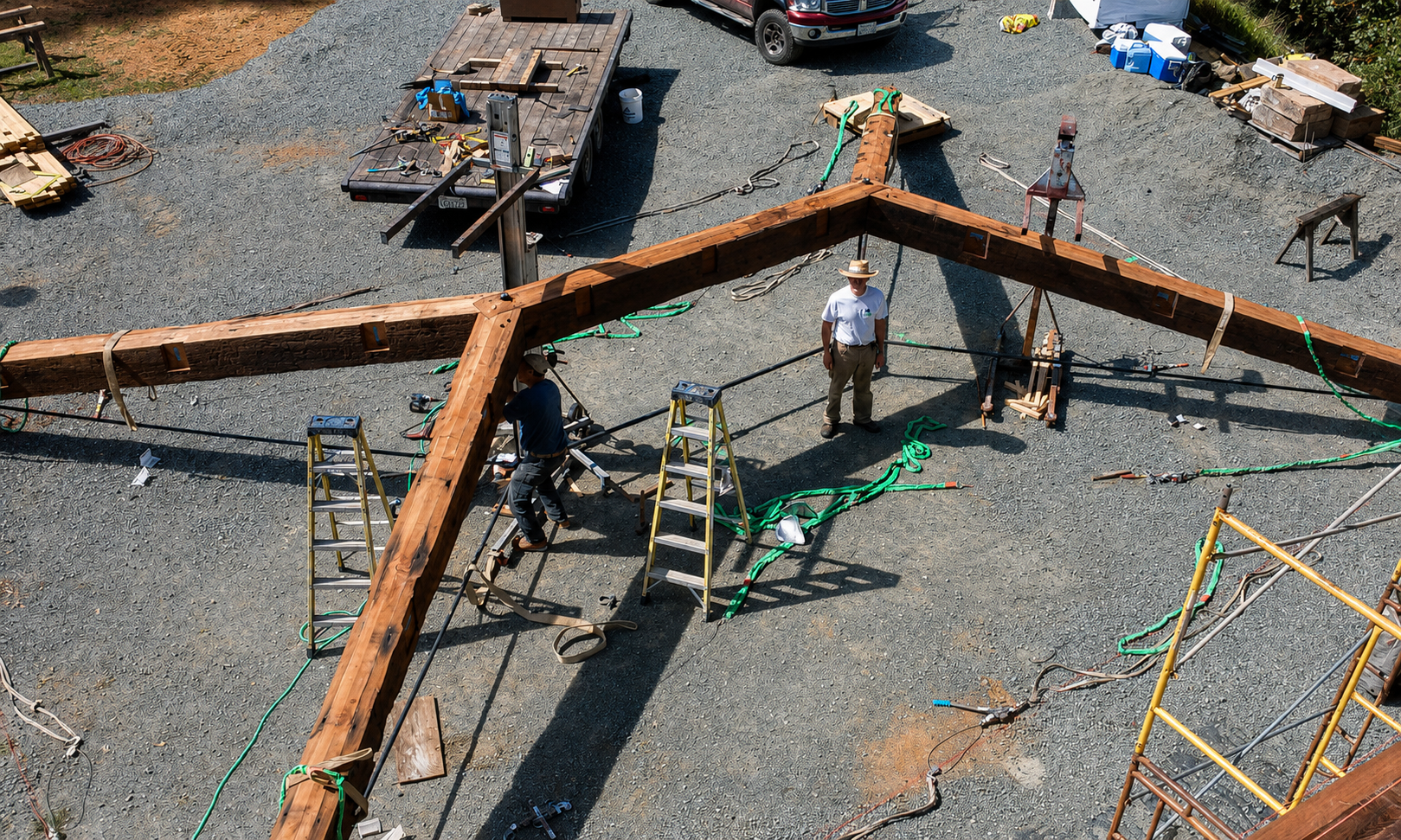Construction site with workers assembling a large wooden beam structure, surrounded by tools, scaffolding, ladders, and vehicles on a gravel lot.