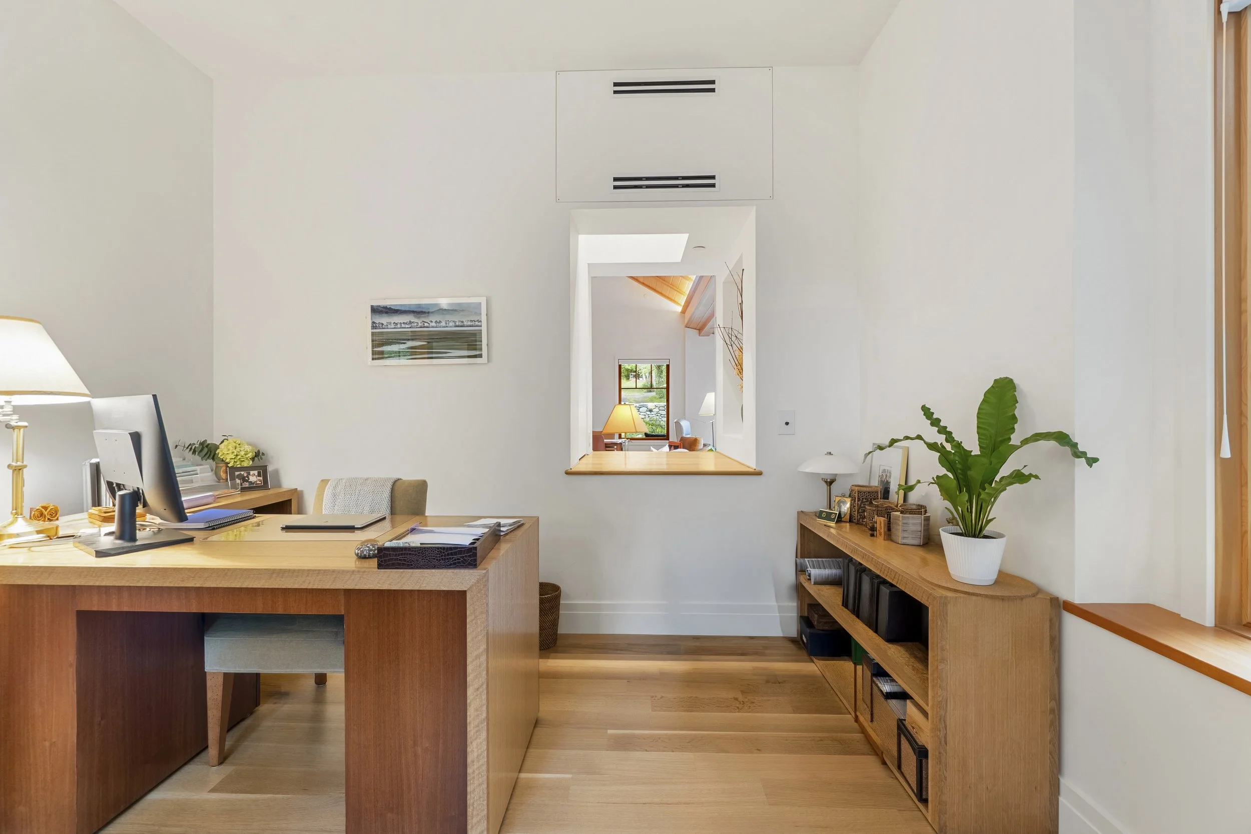 A modern home office with a large wooden desk, a computer, books, and decorative items. There is a window in the background, a potted plant, and artwork on the walls. The decor is minimalist with warm wood tones and white walls.