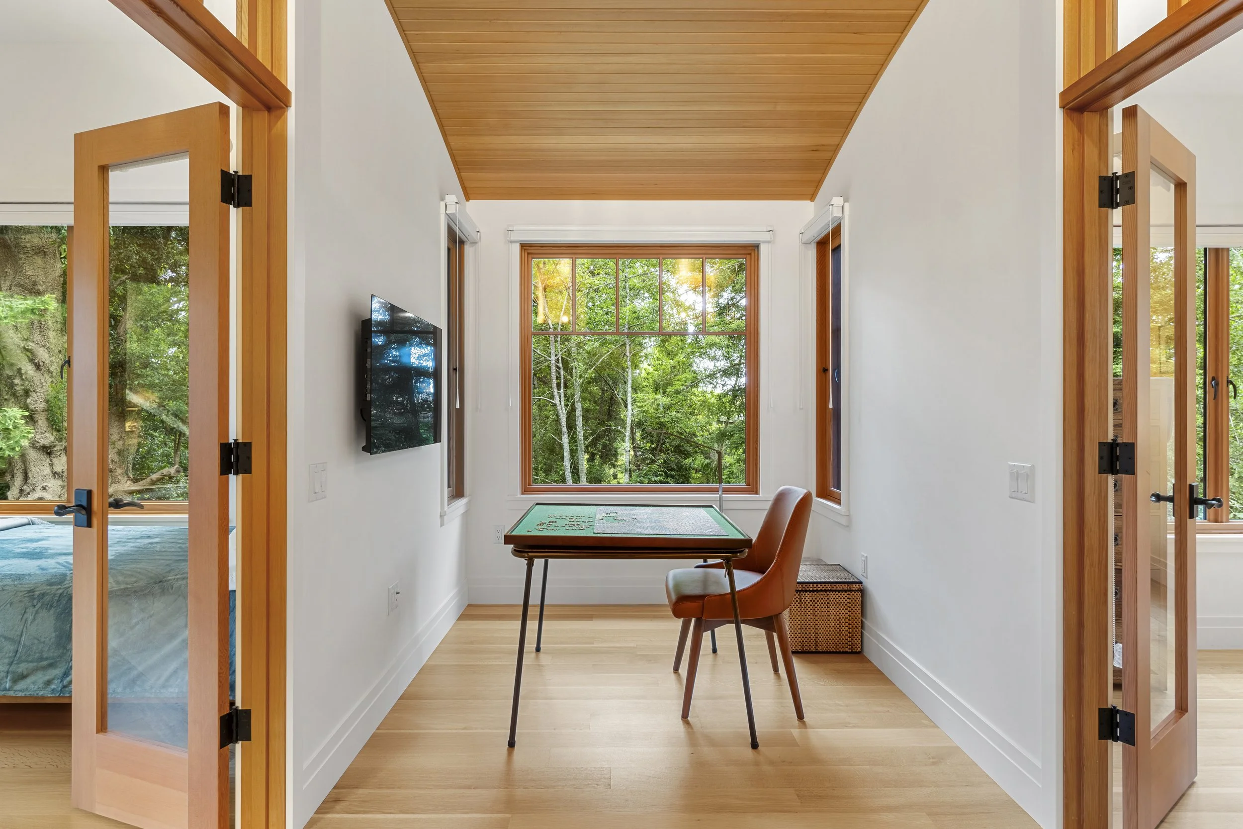 A cozy room with white walls and natural wood ceiling, featuring large windows showing green trees outside. In the room, there is a small table with puzzles and a chair, with a wall-mounted TV nearby.