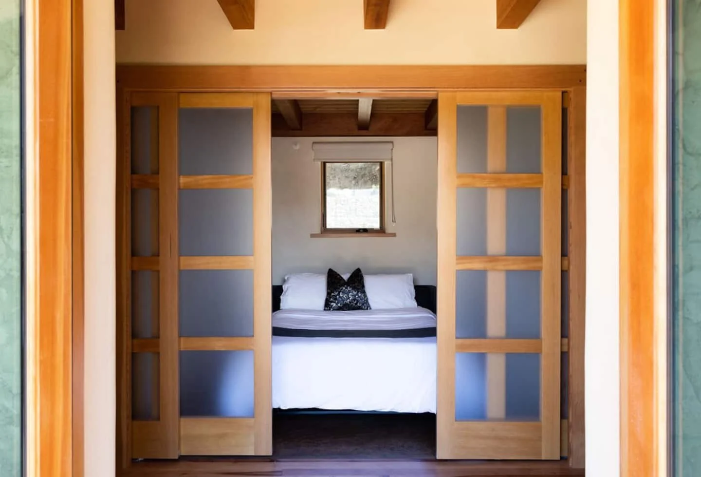 View through sliding wooden doors into a bedroom with a bed, black and white pillows, and a window with a white blind.