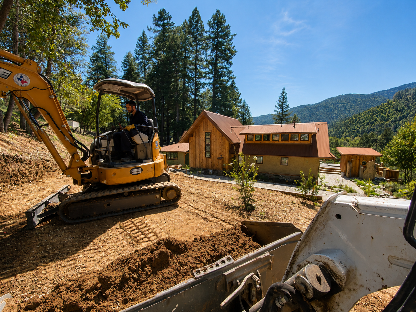 A man operating a small yellow excavator on a dirt hill with a wooden house and trees in the background