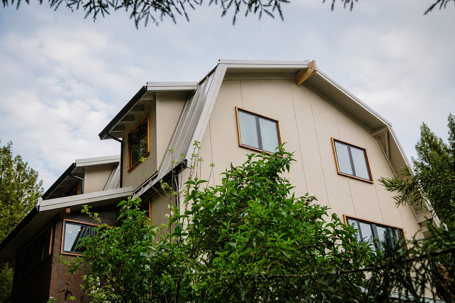 A modern house with multiple windows, metal roofing, and beige exterior walls, surrounded by green foliage, under a partly cloudy sky.