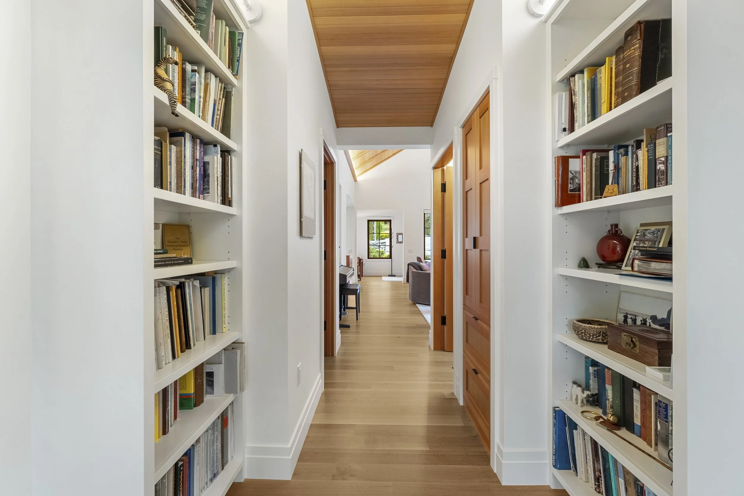A hallway with white walls and wooden floors, flanked by white bookshelves filled with books and decorative items, leading to a living area with large windows and wooden ceiling panels.