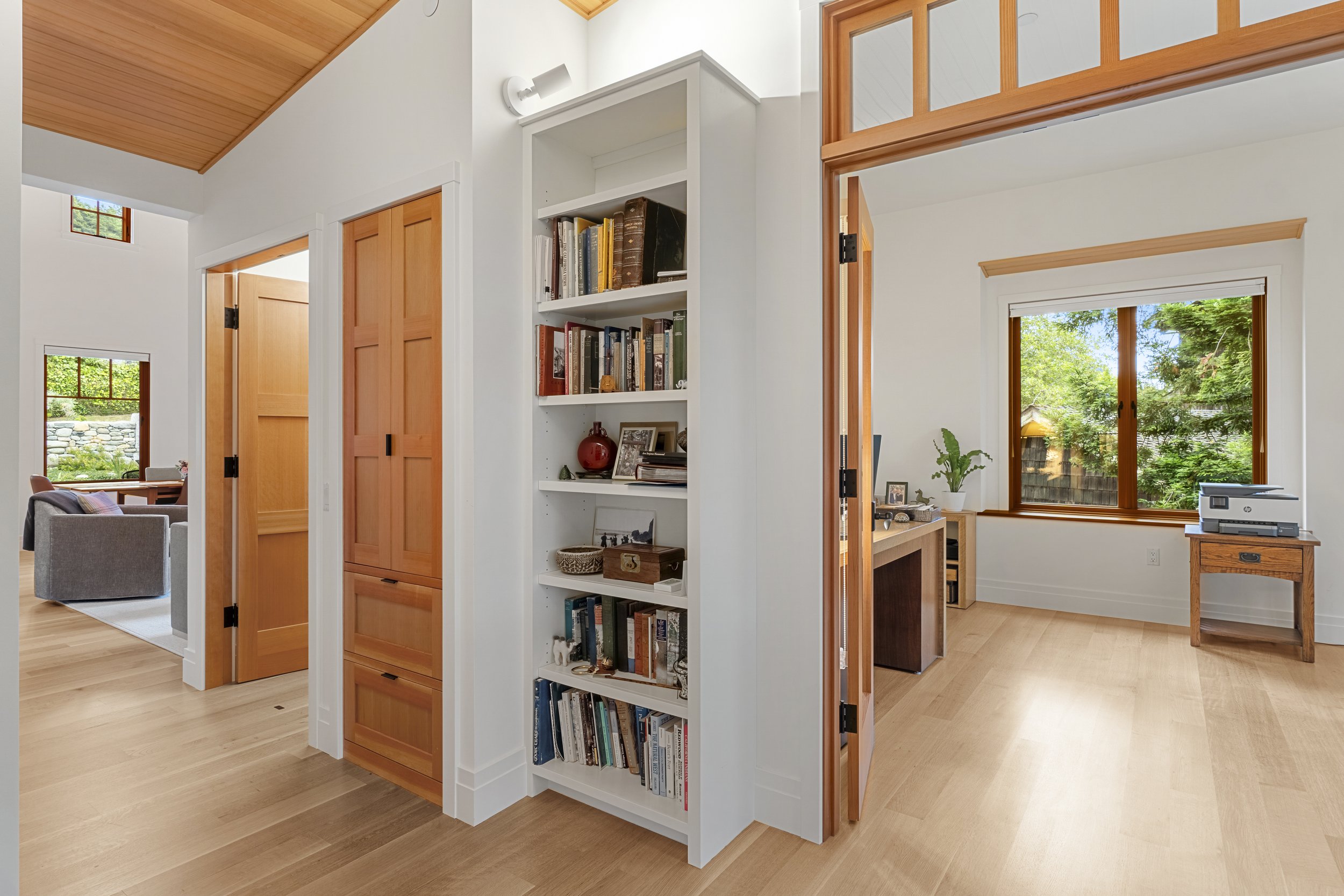 Interior view of a modern home with wooden flooring, white walls, large windows, and built-in shelving filled with books and decorative items. The image shows part of a living space, an open doorway to another room with a desk and window, and a partl