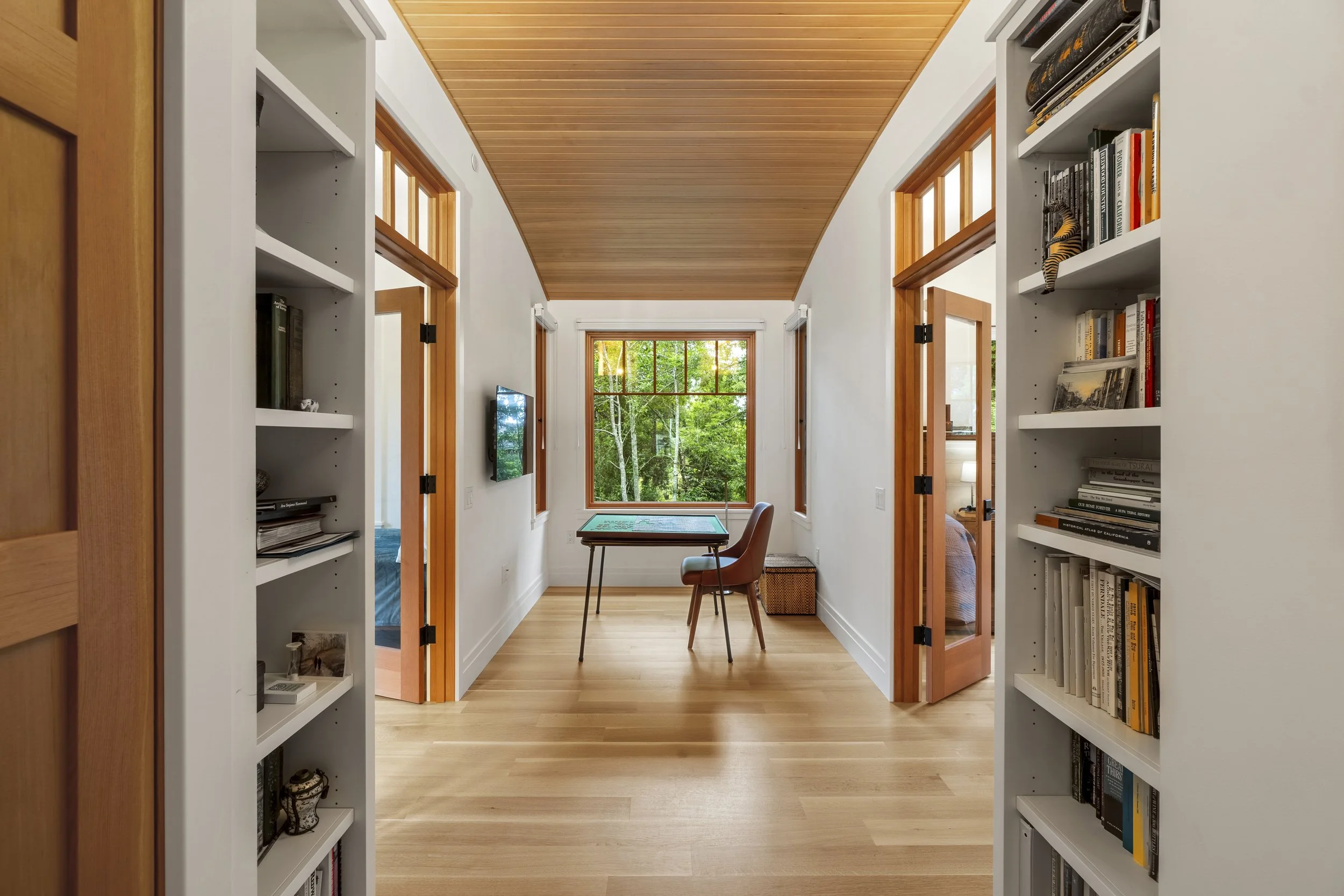 Interior view of a cozy room with large windows showing a lush green outdoor landscape, a small table and a chair in front of the window, surrounded by white bookshelves filled with books, and a warm wooden ceiling.