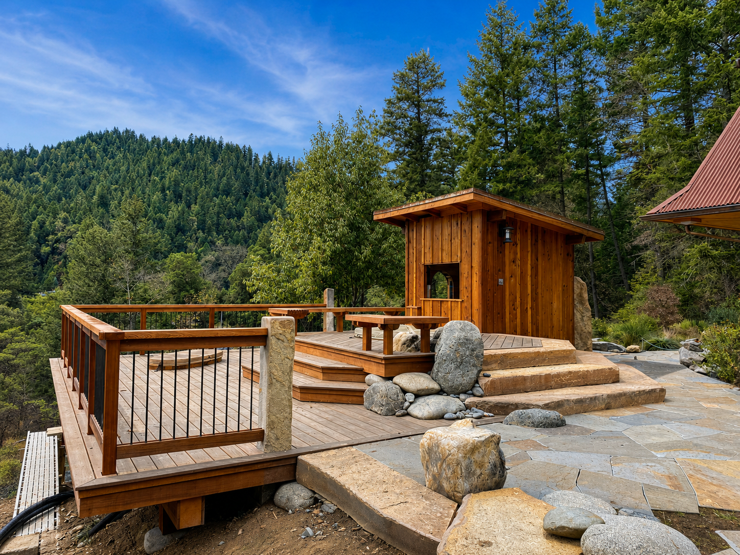 A wooden deck with a small wooden structure and outdoor furniture, surrounded by rocks and trees, overlooking a lush forested hillside under a blue sky.