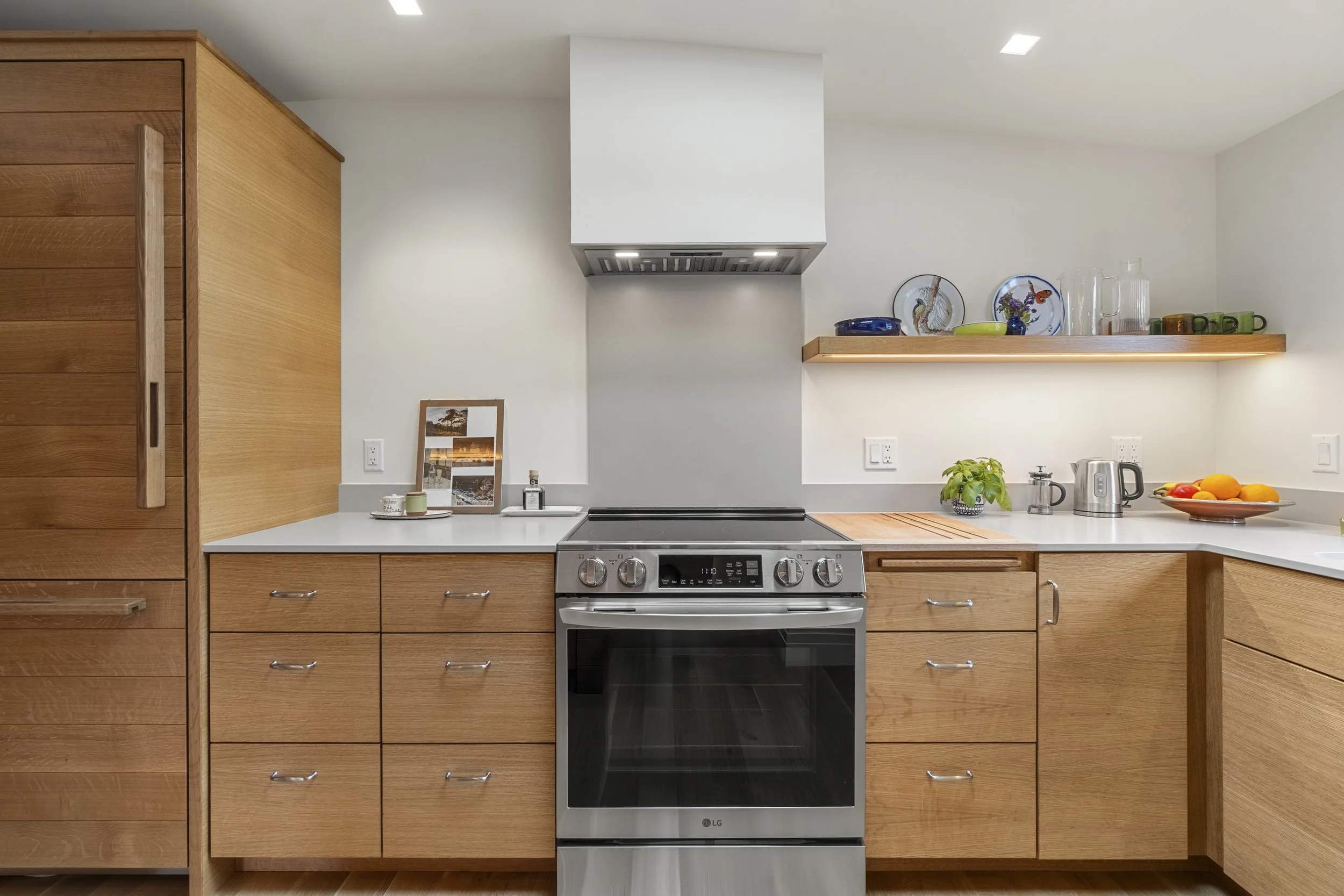 Modern kitchen with wooden cabinets, a stainless steel oven, a white counter, and a shelf with decorative plates, glasses, and cups.