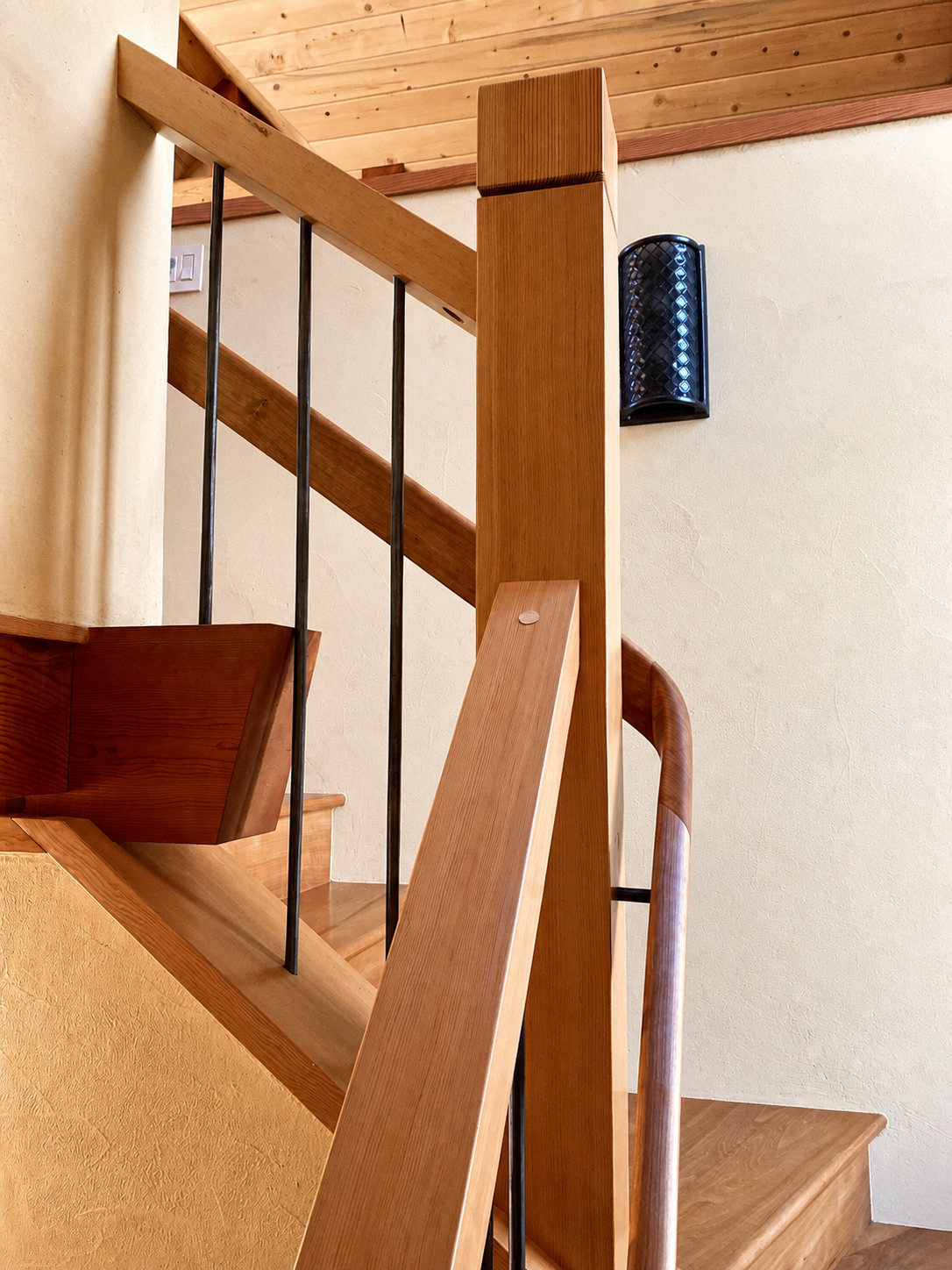 Close-up view of wooden staircase with metal balusters, interior wall with wall-mounted black light fixture, wood-paneled ceiling, and light-colored walls.