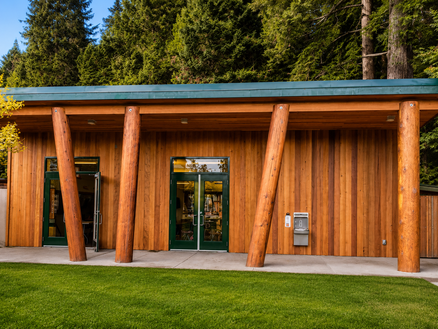 Modern building with vertical wooden siding, green glass double doors, large angled wooden support beams, and surrounded by a grassy lawn with trees in the background.