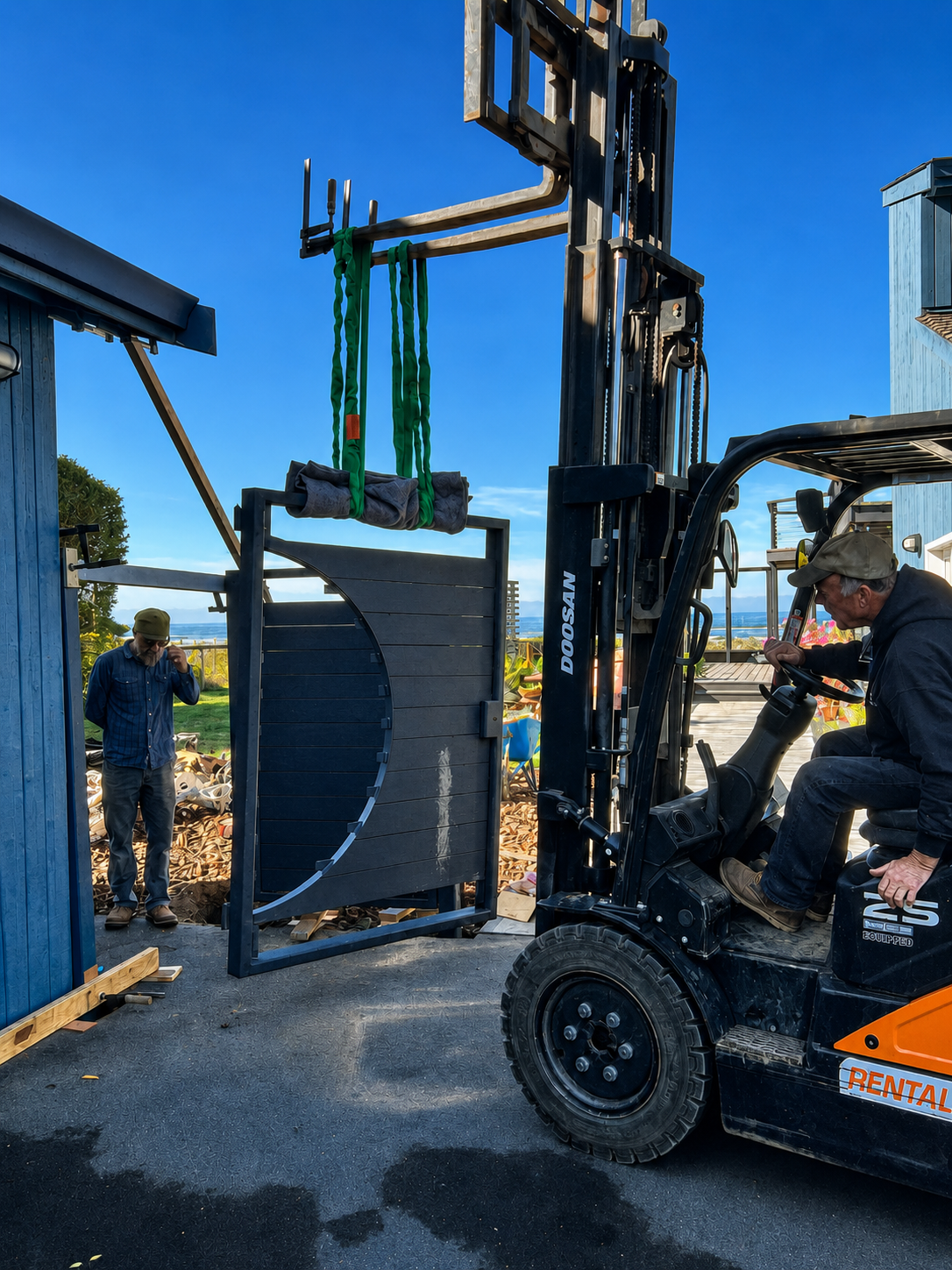 Two men working outside, one operating a forklift lifting a large dark-colored structure, the other standing nearby, with a blue building and a bright blue sky in the background.