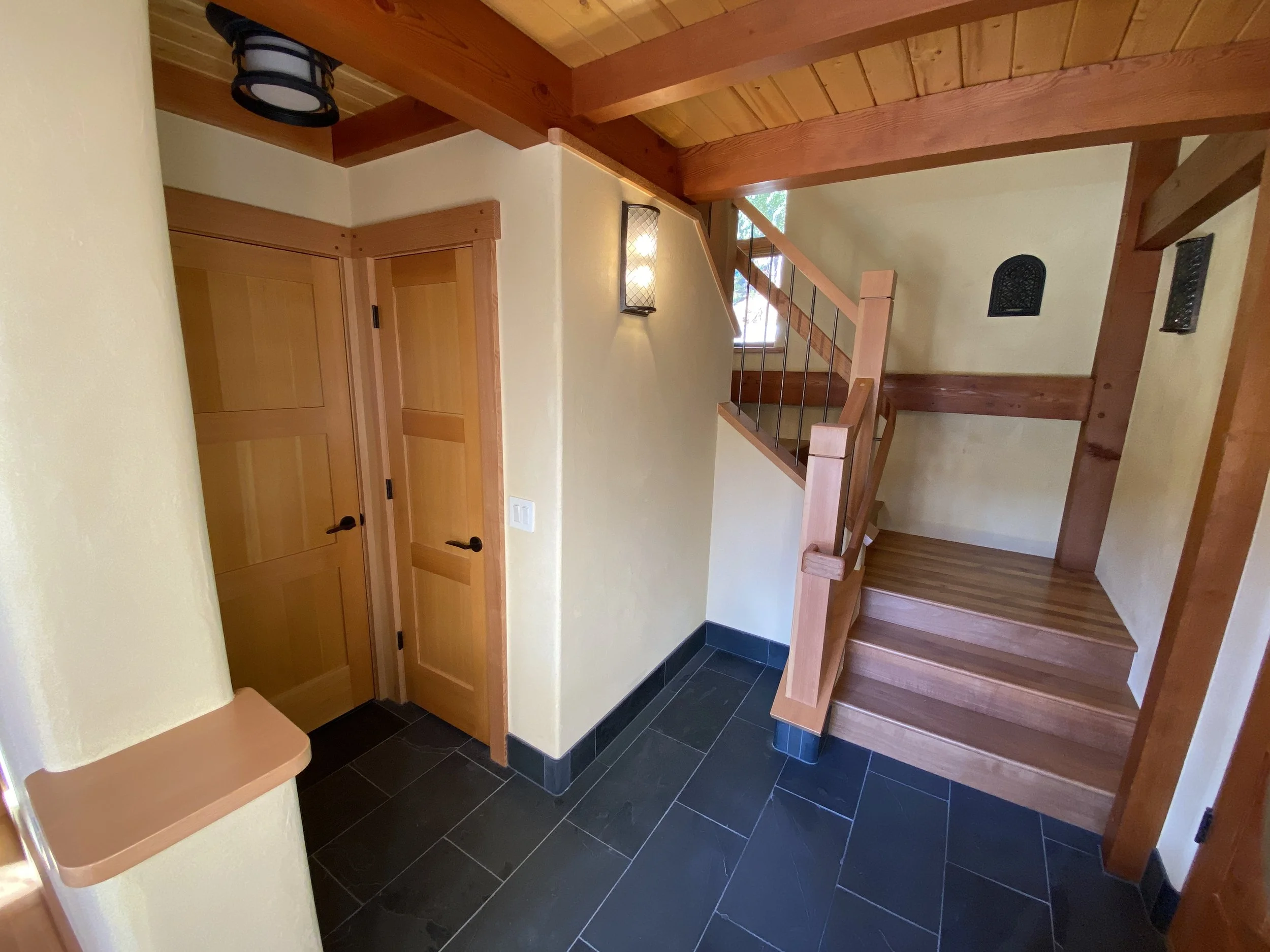 Interior of a home with wooden accents, including a staircase with wooden steps and railing, dark tile flooring, and yellow walls. There are two black wall-mounted decorative grilles and a window that lets in natural light.