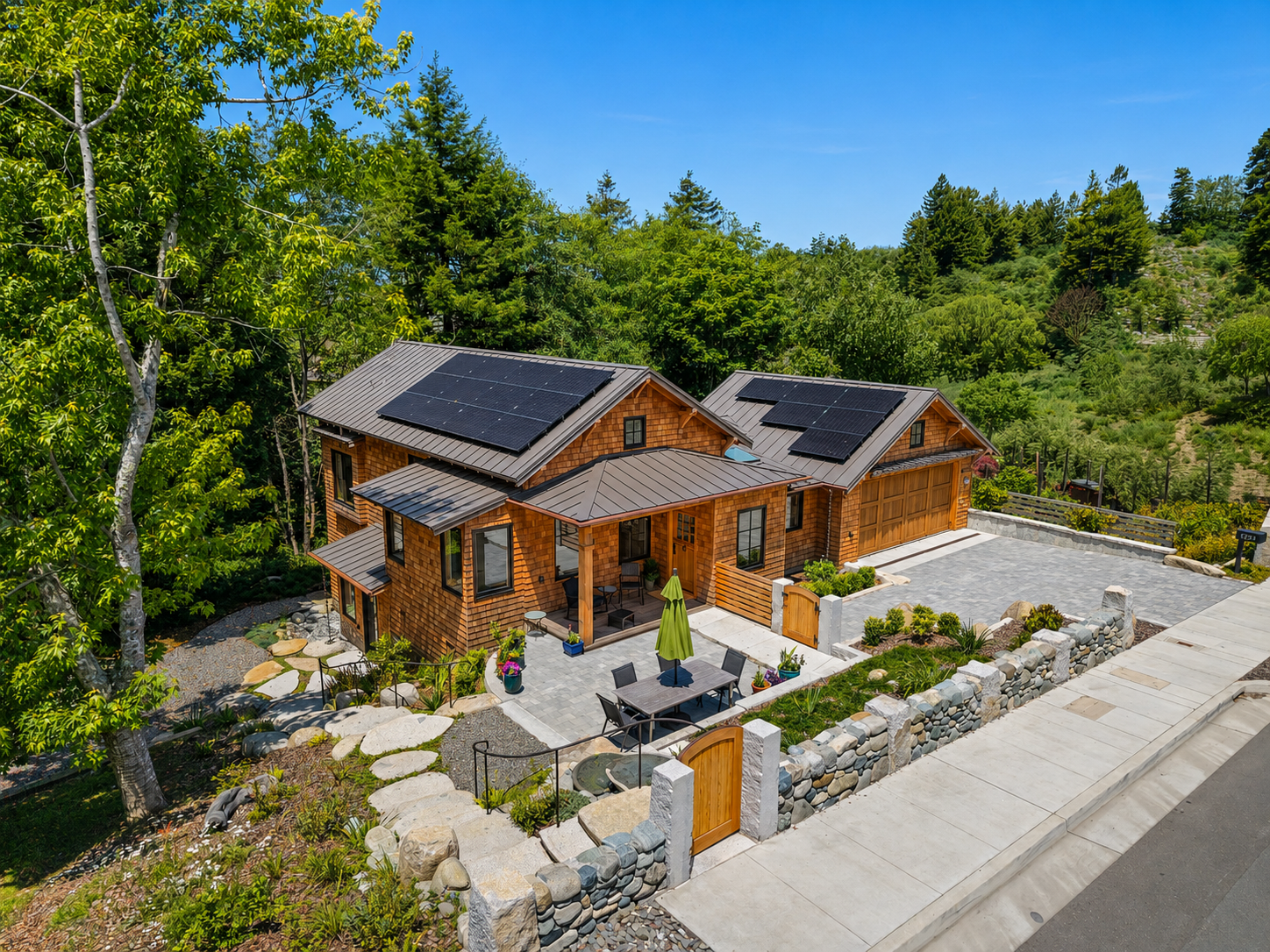 A house with a metal roof covered with solar panels, surrounded by green trees and a garden with outdoor furniture, in a hilly landscape.