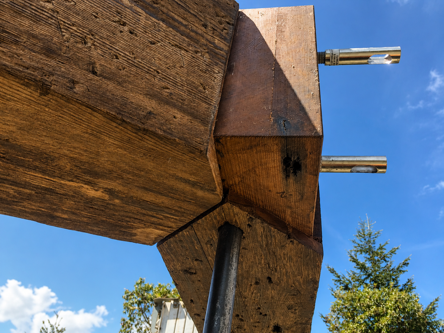 Close-up of a wooden structure with metal rods extending from it, set against a blue sky with some clouds and trees in the background.