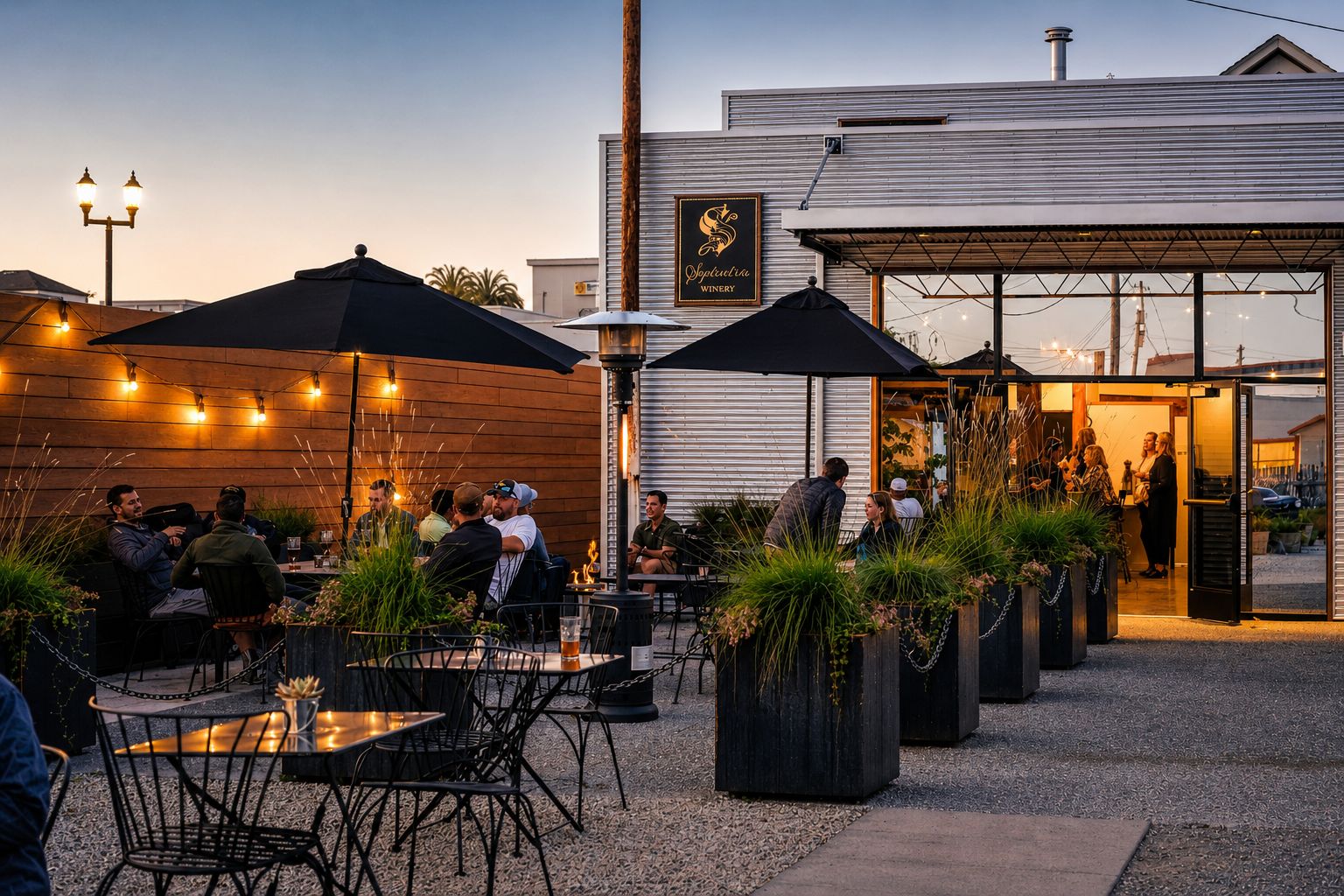 Outdoor dining area of a winery with people enjoying evening drinks, decorated with string lights, large umbrellas, and lush plants, with a modern building in the background.