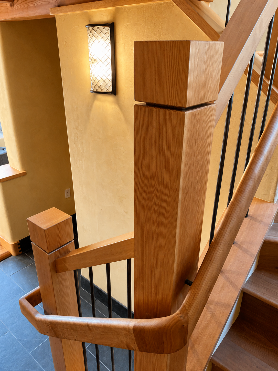 Close-up view of a wooden staircase railing and newel post with black metal balusters, in a warmly lit interior space.