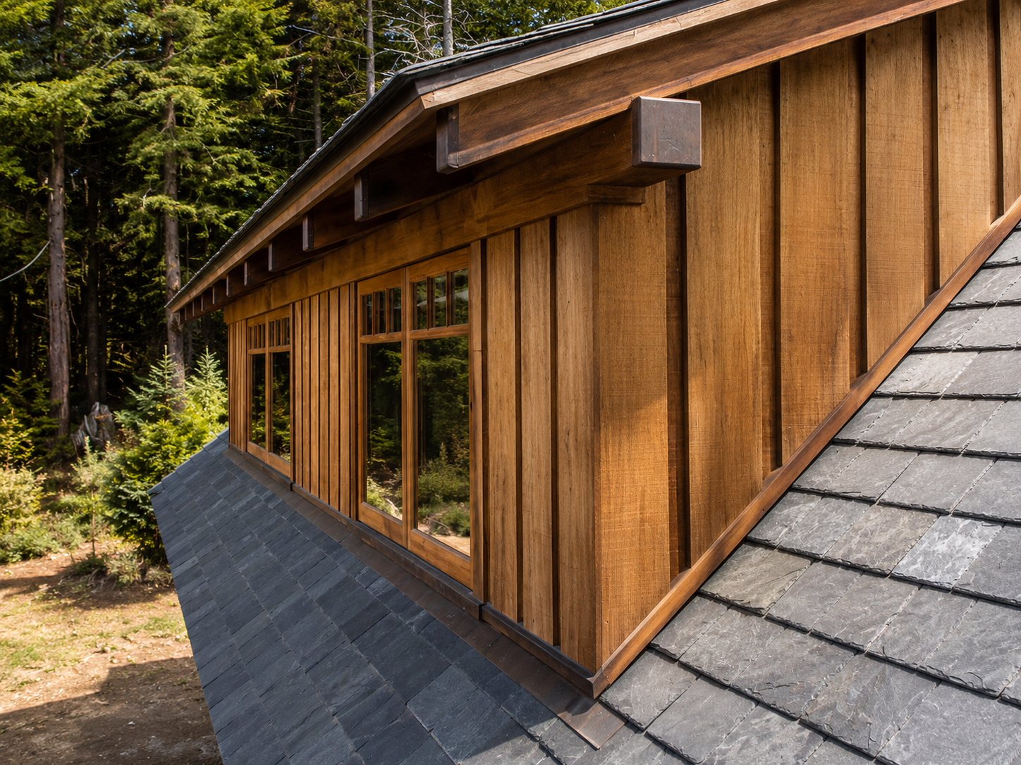 Close-up of a wooden house with large windows, set against a green forest background. The house has vertical wooden siding and a sloped roof with gray shingles.