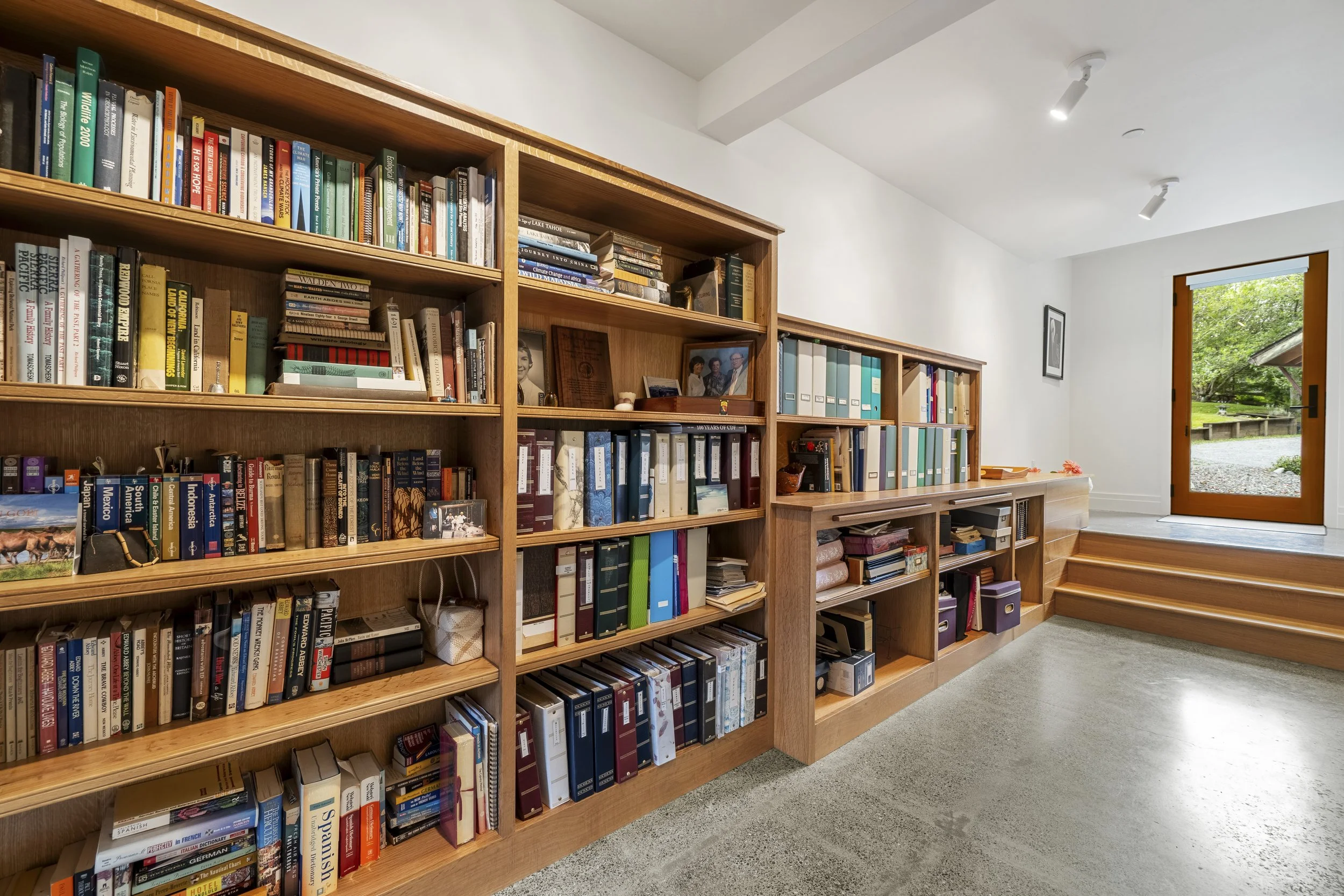 A wooden bookshelf filled with various books, some binders, and framed photographs sits against a white wall in a bright room. An open door with a view of greenery outside is on the right side of the image.