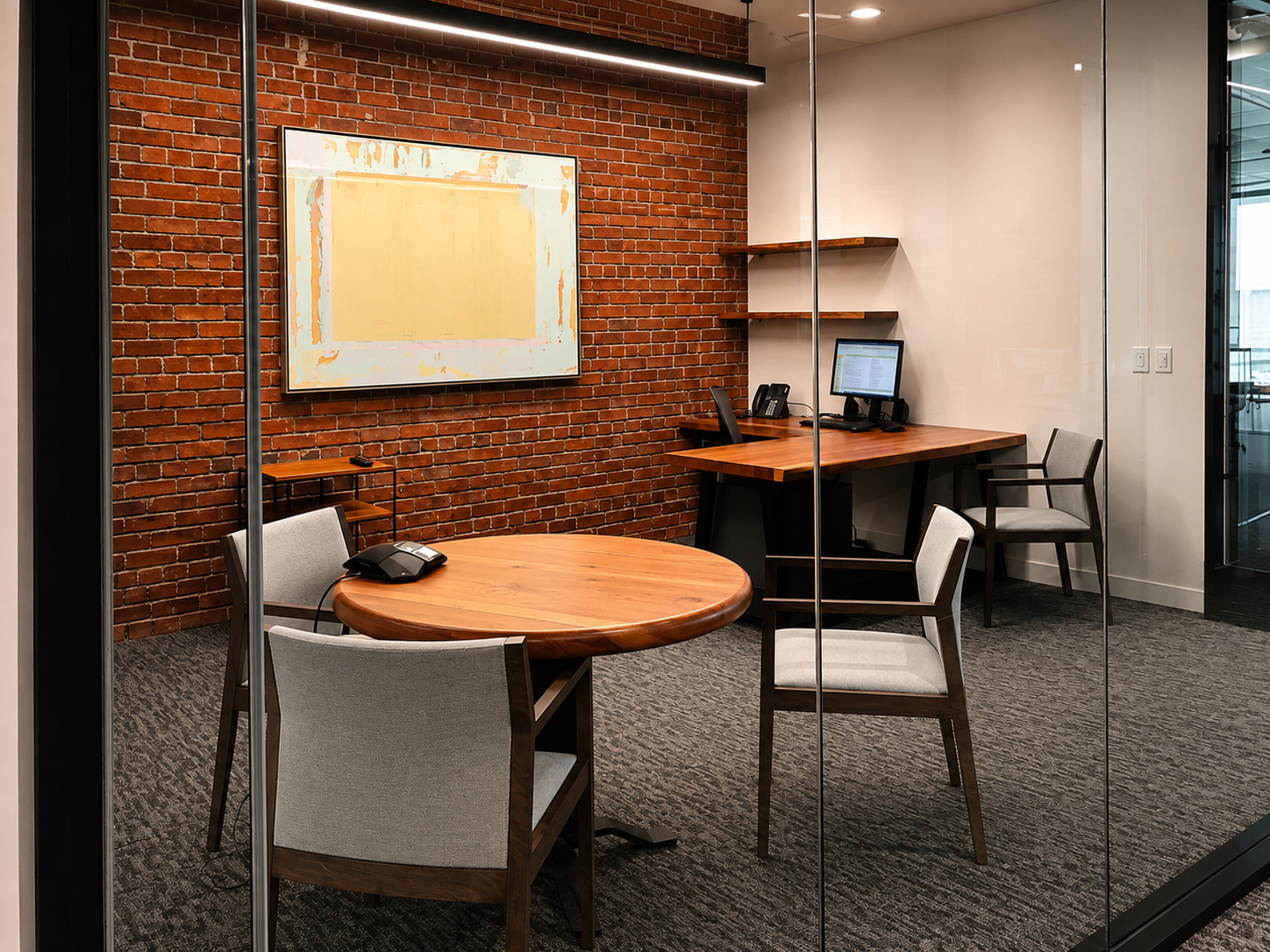 An office conference room with a brick accent wall, a round wooden table with four chairs, a rectangular desk with a computer and phone, and a large wall-mounted screen.