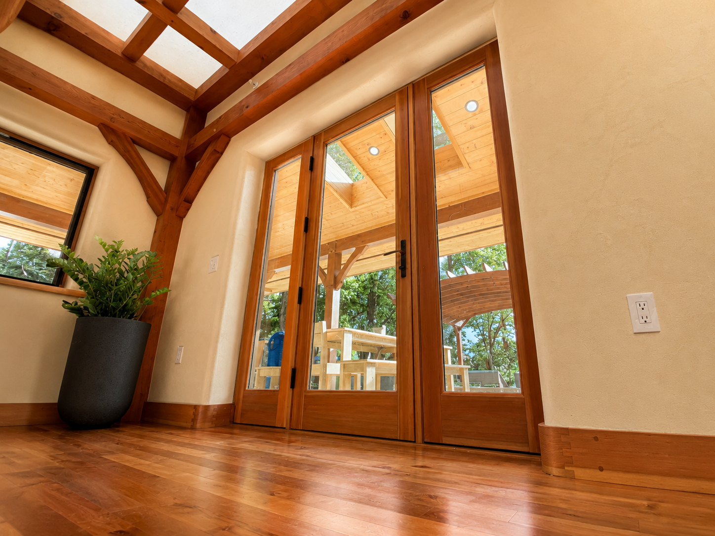 Interior view of a room with wooden floors, large glass sliding doors, a potted plant, and exposed wooden beams.