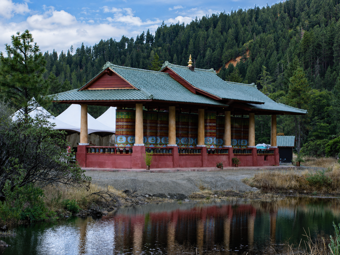 A traditional Buddhist temple with a blue tiled roof, red walls, and decorative prayer wheels, set against a forested mountain landscape and reflected in a small pond.
