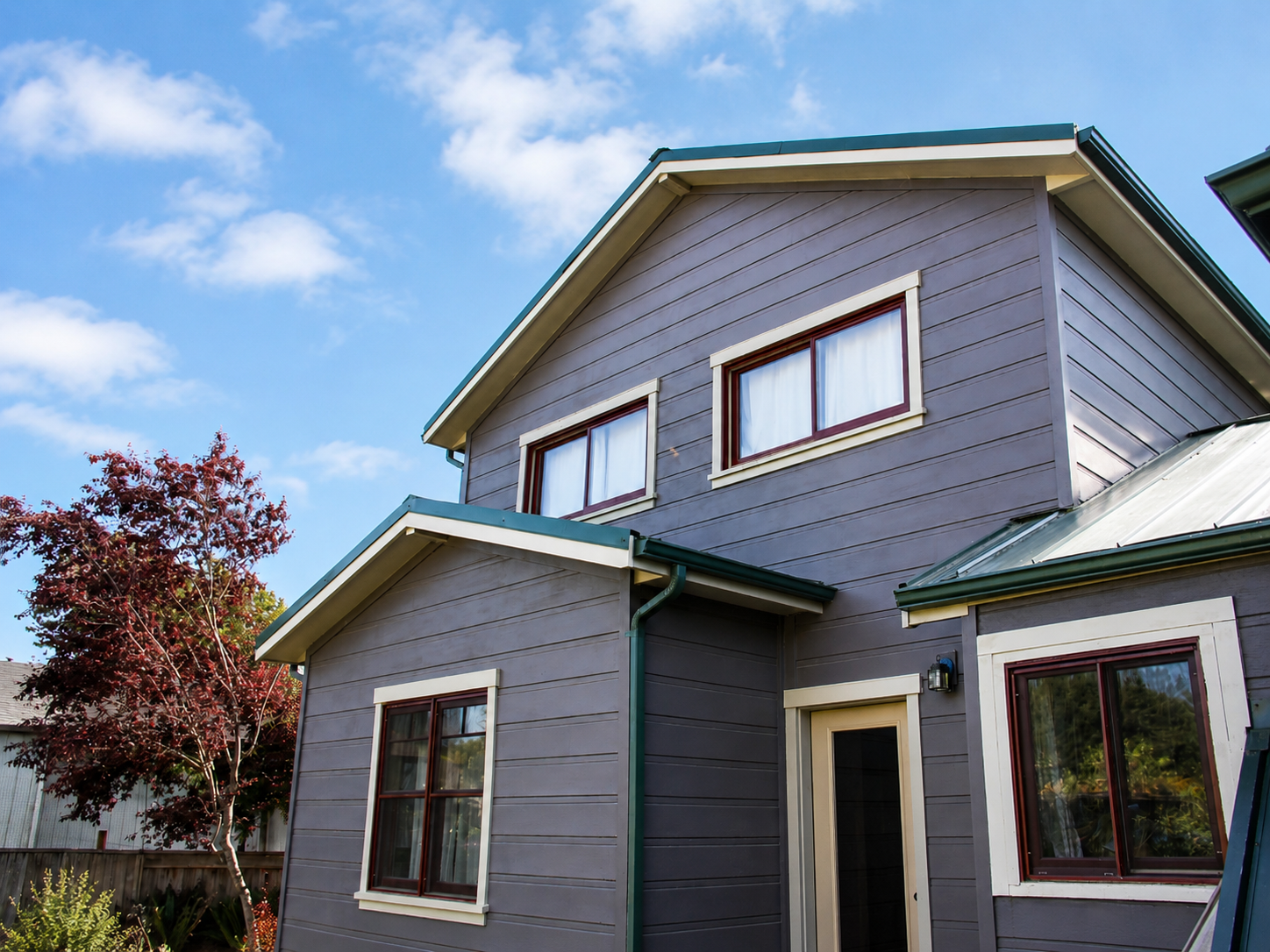 A two-story house with gray wooden exterior and green roof, featuring multiple white-framed windows, a small side entrance, and a tree with red leaves in the yard under a partly cloudy blue sky.