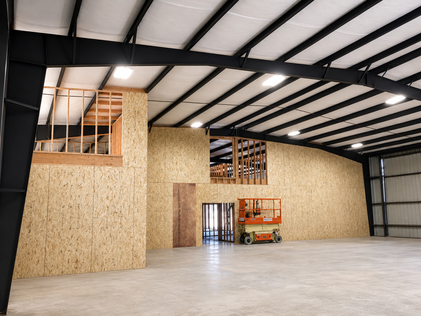 Interior of a building under construction with unfinished wooden walls, a lift, and unfinished second story with railing.