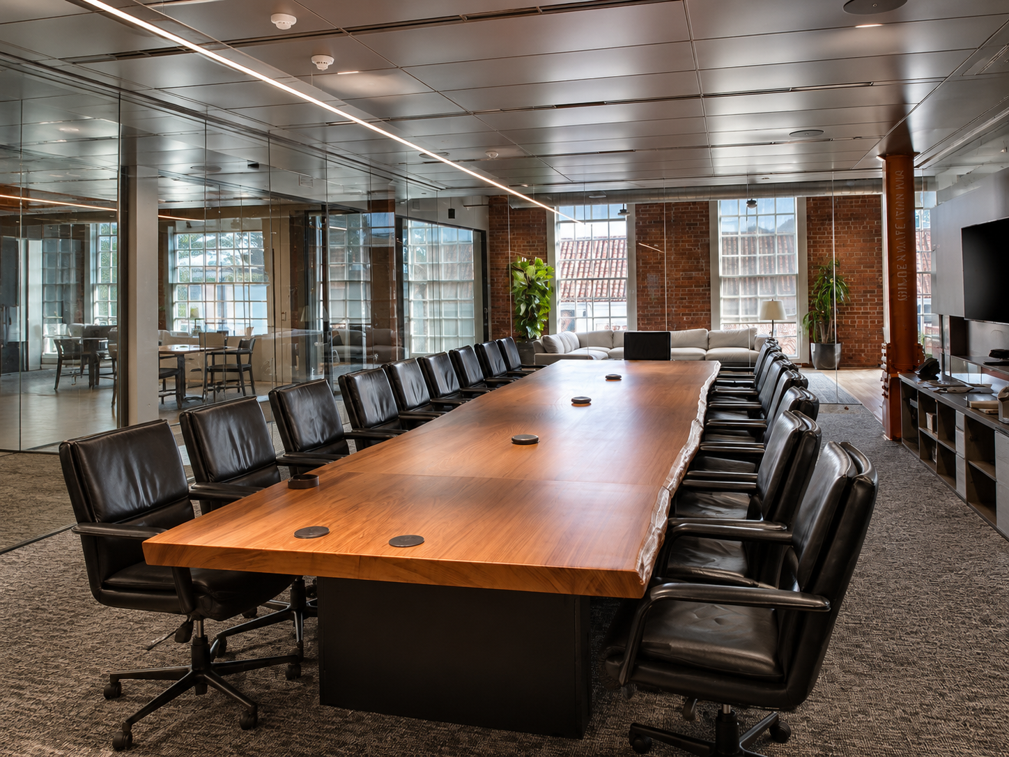 A modern conference room with a large wooden table surrounded by black leather chairs, located in an office with glass walls, large windows, and exposed brick walls behind a seating area with a white sofa and plants.