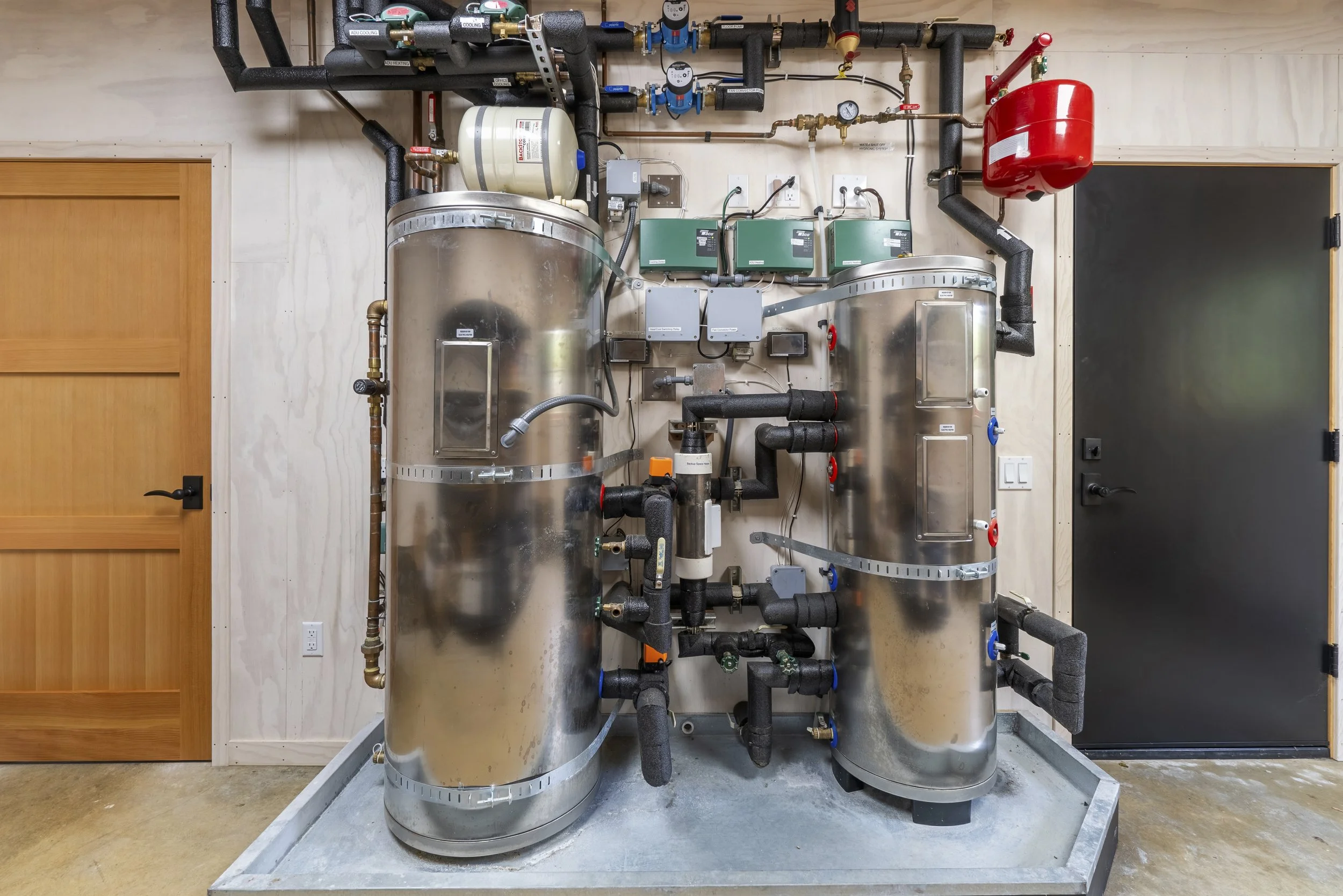 A mechanical system with large stainless steel tanks, black pipes, gauges, electrical boxes, and control valves in a utility room.