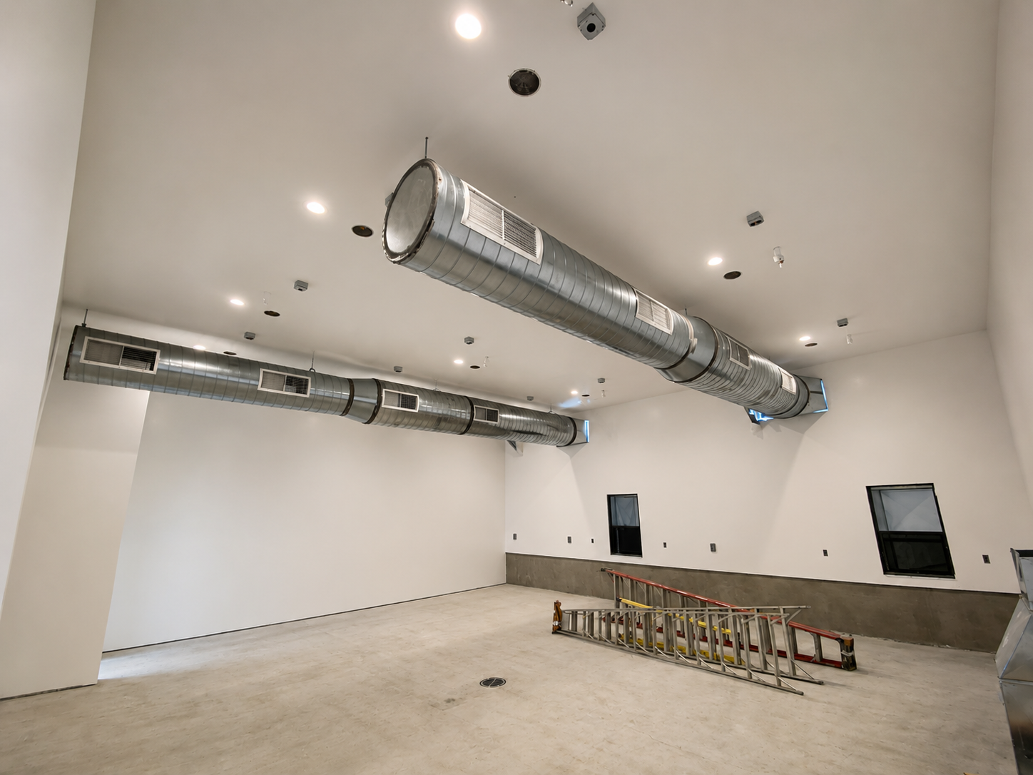 Empty room with exposed industrial HVAC ductwork on the ceiling, ladder on the floor, and small windows along the wall.