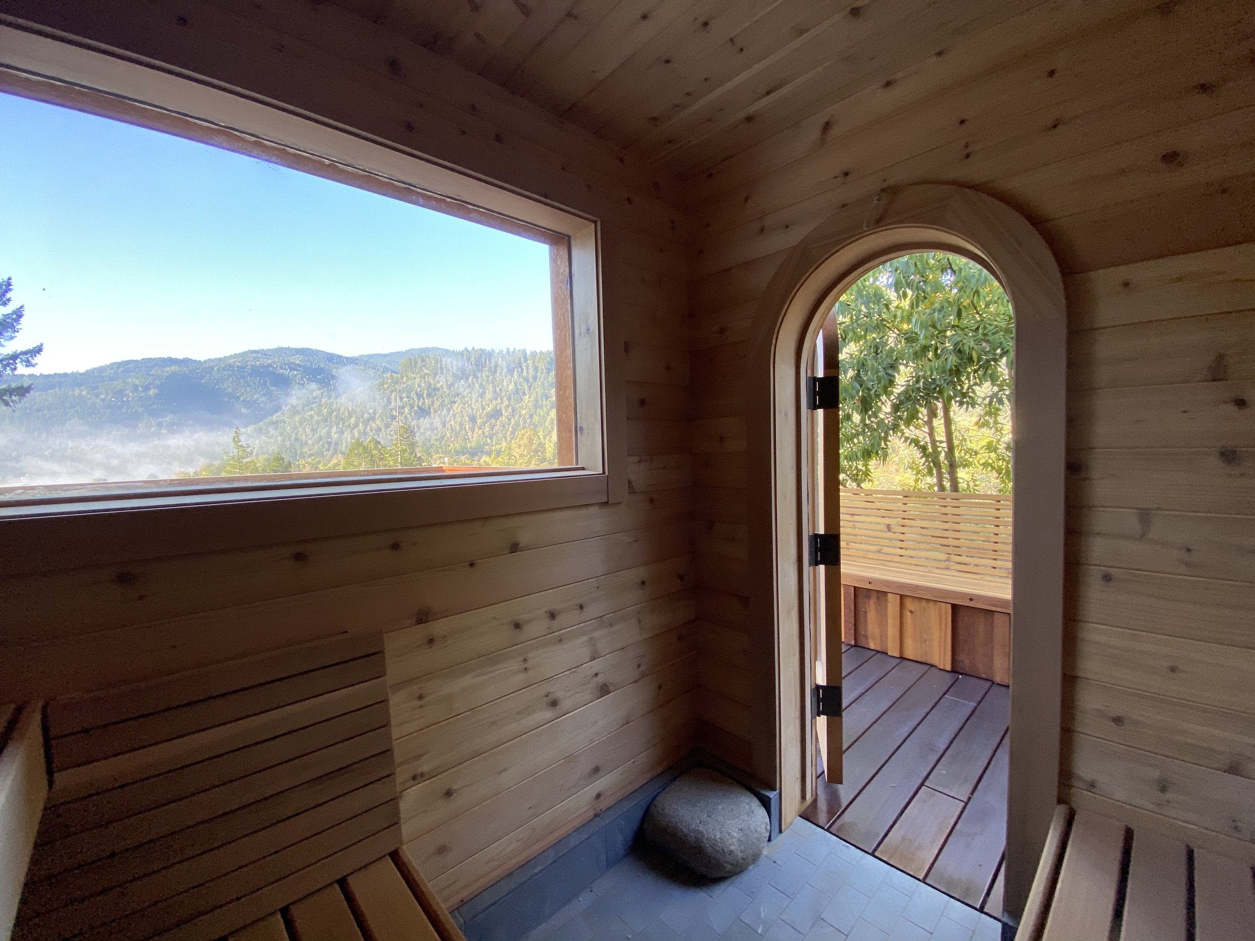 Wood-paneled interior with a rectangular window showing a mountain landscape and a small arched doorway leading outside where trees and a wooden fence are visible.