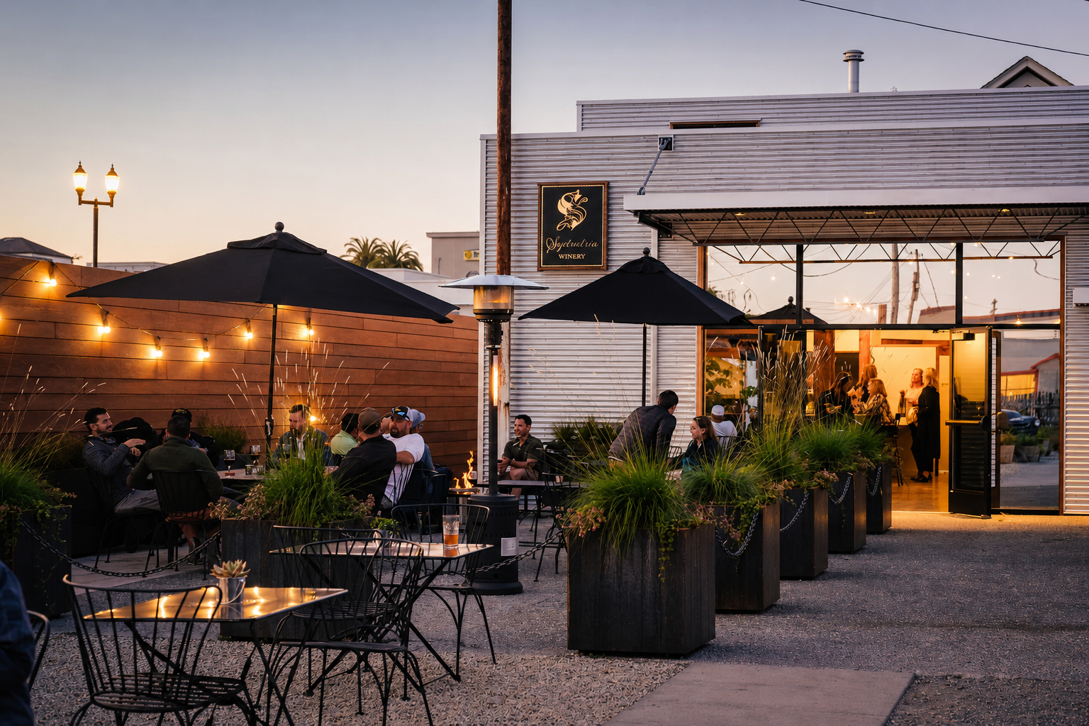 People dining and socializing outside at a winery during the evening with string lights, large umbrellas, and a fire pit, in front of a modern building with a winery sign.