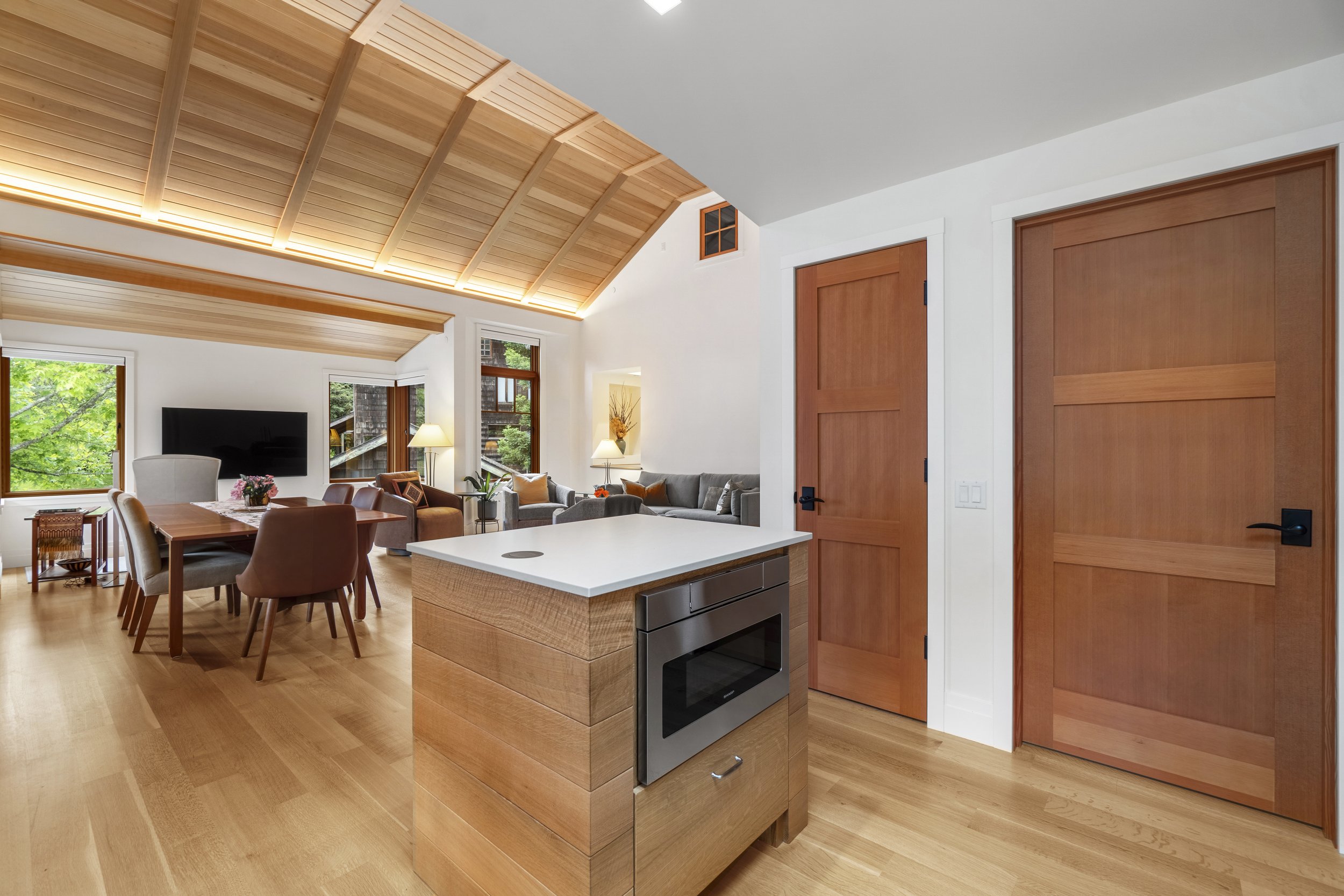 Open-concept living room and kitchen area with vaulted wooden ceiling, large windows, hardwood floors, and modern furniture, including a gray sofa, dining table, and a kitchen island with an oven.