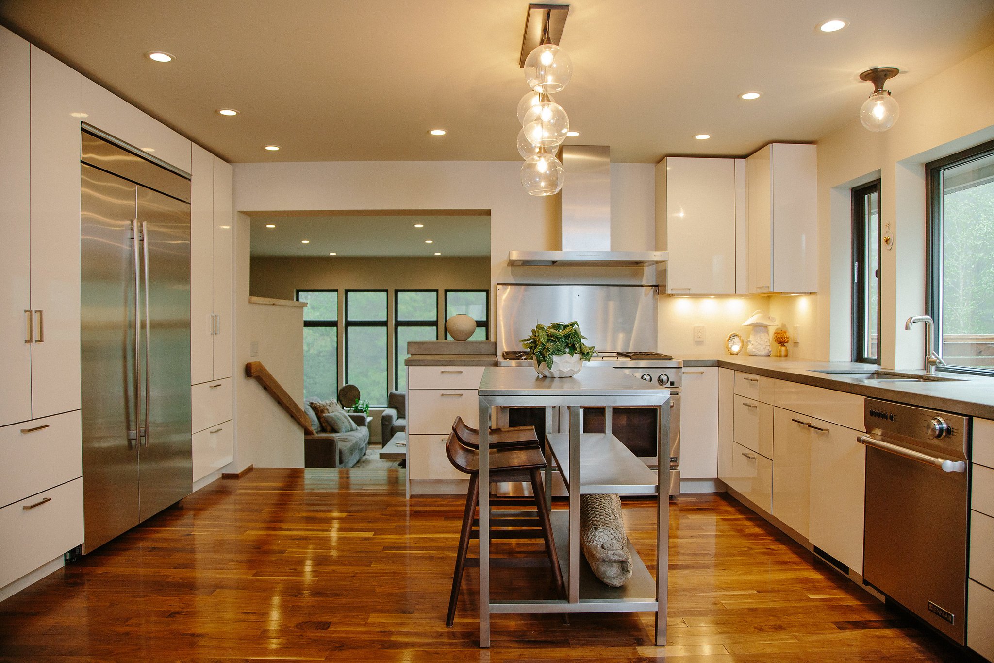 Modern kitchen with white cabinets, stainless steel appliances, pendant lights, large windows, hardwood floors, and a kitchen island with seating.