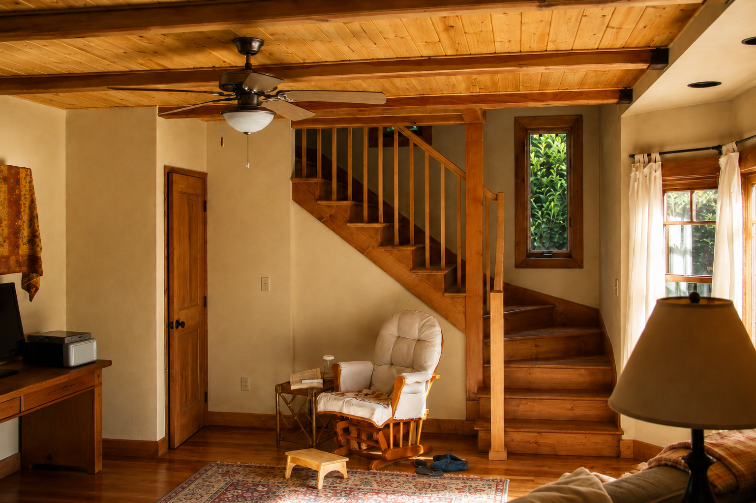 Living room with wooden stairs, a beige armchair, a window with curtains, a ceiling fan, and a wooden cabinet.