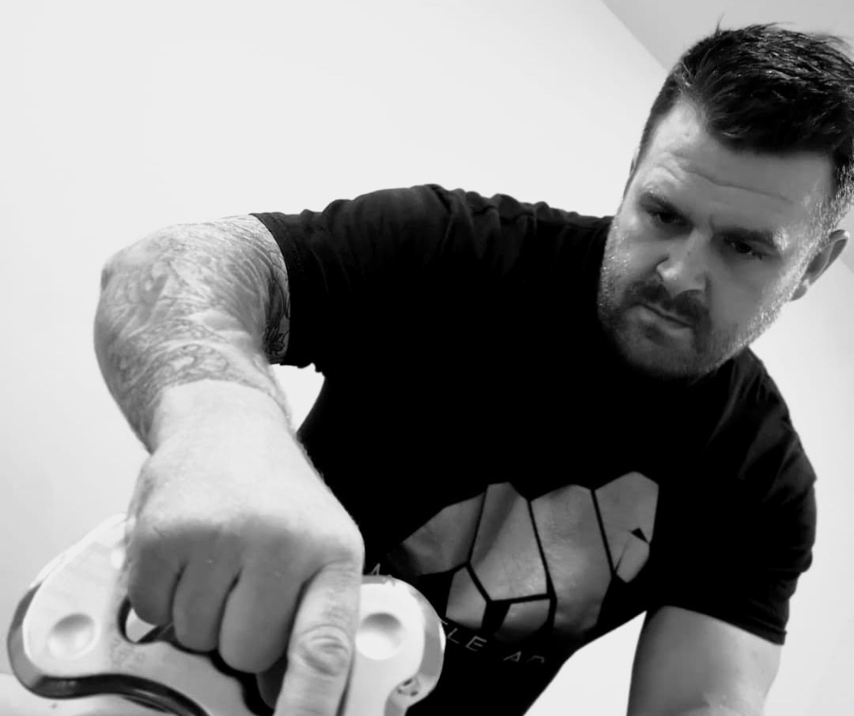 A man with tattoos on his arm, wearing a black T-shirt, appears to be grabbing or holding a gaming controller or device, with a serious expression on his face.