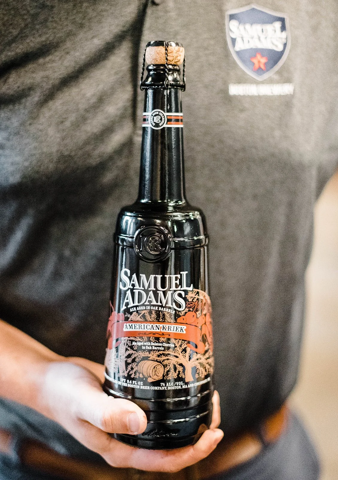 Person holding a beer bottle labeled Samuel Adams American Kriek, dark bottle with decorative label, person wearing a gray shirt with Samuel Adams logo.