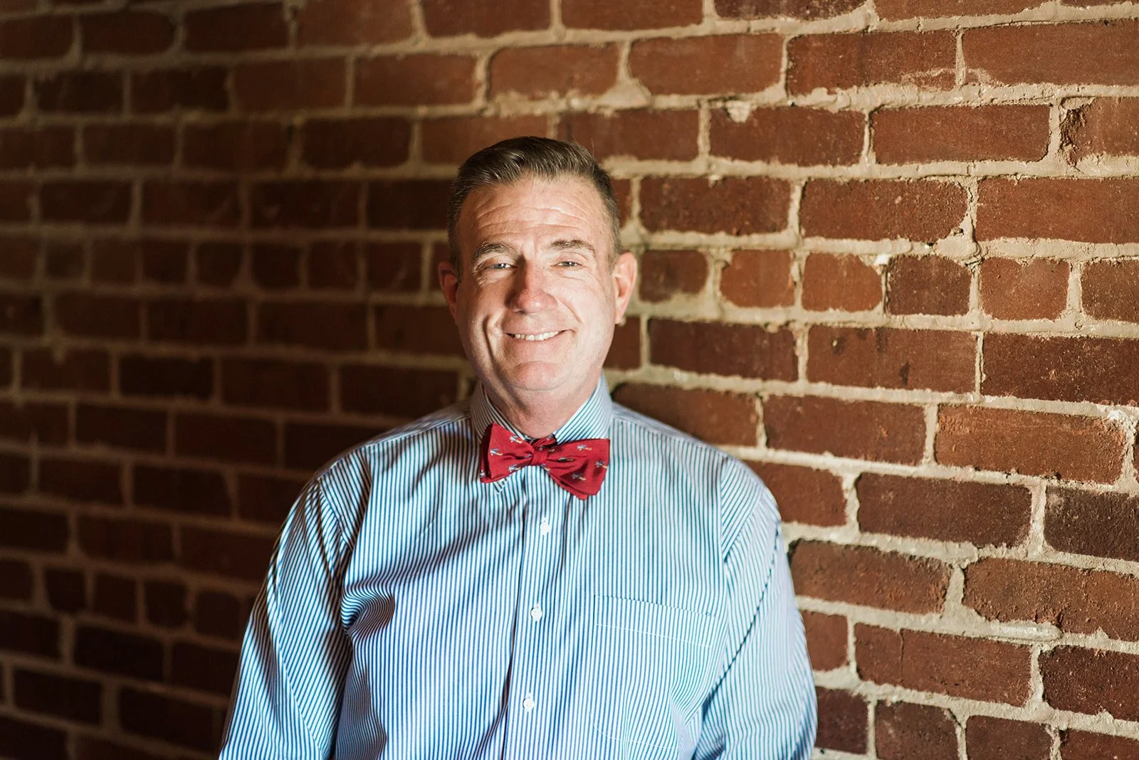 A smiling man wearing a blue striped shirt and a red bow tie, standing against a brick wall.