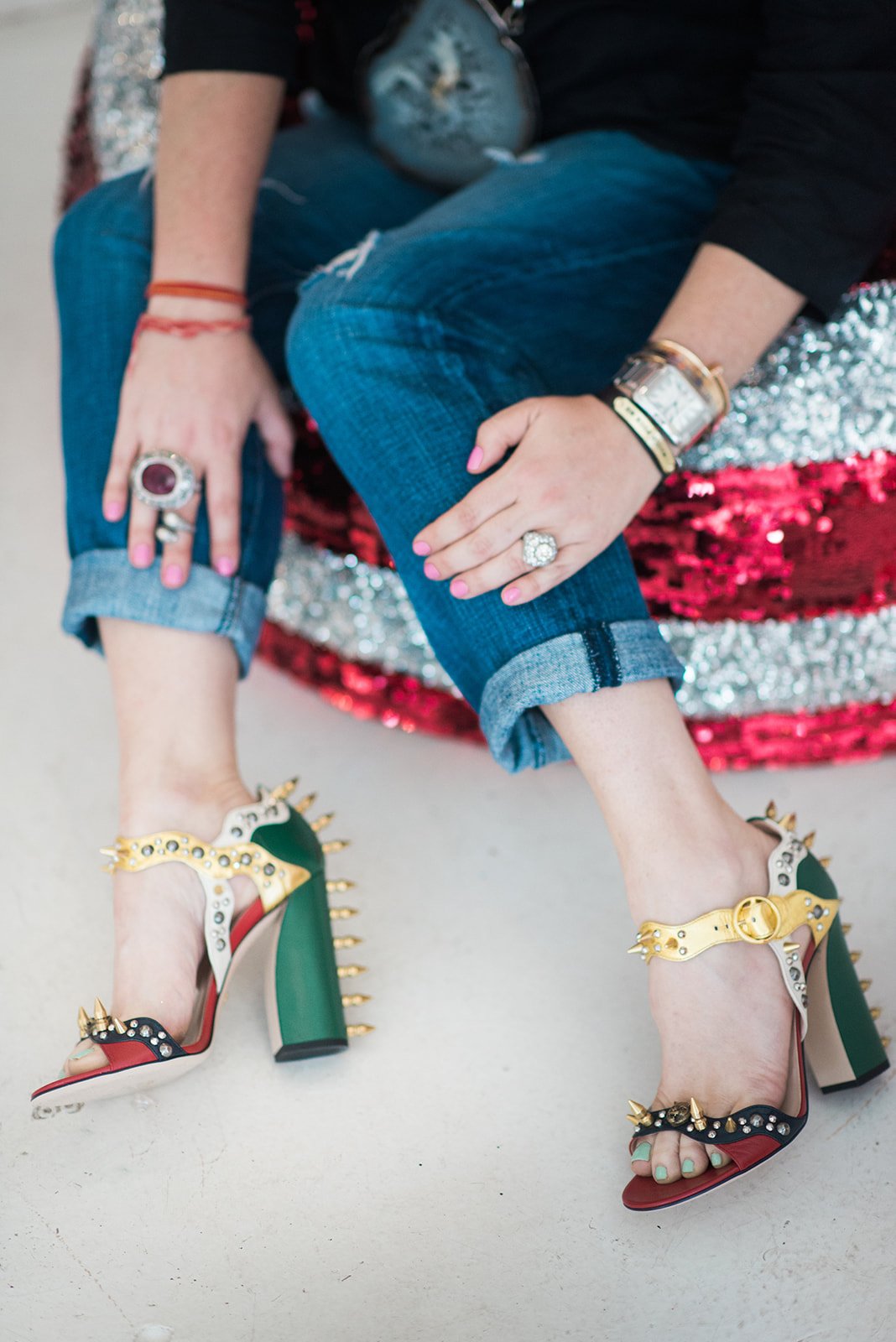 A person is sitting on a sparkly red and silver sequin-covered chair, wearing colorful high-heeled sandals with studs and spikes, blue rolled-up jeans, a black jacket, and multiple bracelets and rings.