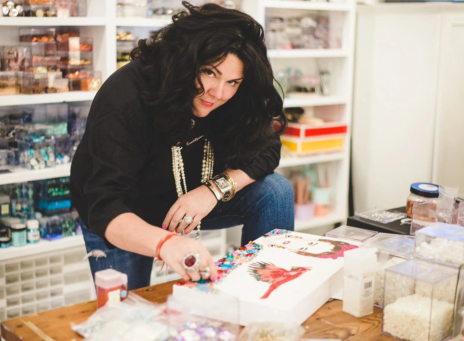 A woman with black curly hair, wearing a black top and jewelry, is working on an art project at a cluttered table in a craft room filled with supplies and organized containers.