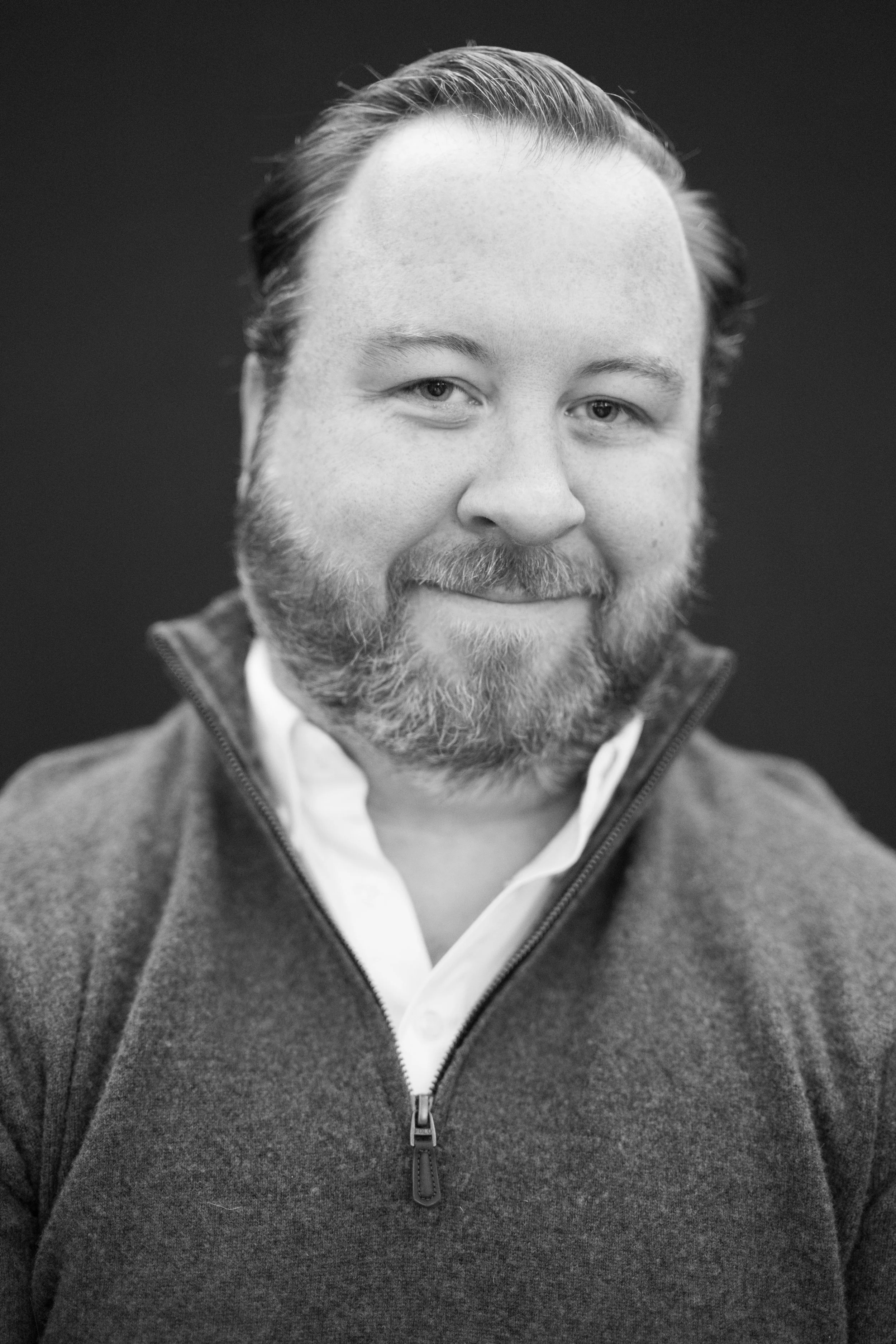 Black and white portrait of a man with a beard, smiling, wearing a zip-up sweater and a collared shirt, against a dark background.