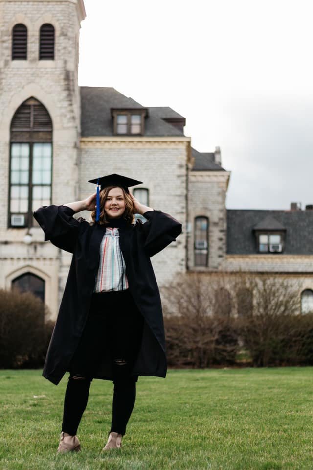 a graduation photo of a woman with dark brown hair and hazel eyes wearing a black graduation cap and gown and a striped shirt, black jeans, and tan leather heels. She's standing outside in front of a large limestone buildings with arched windows.