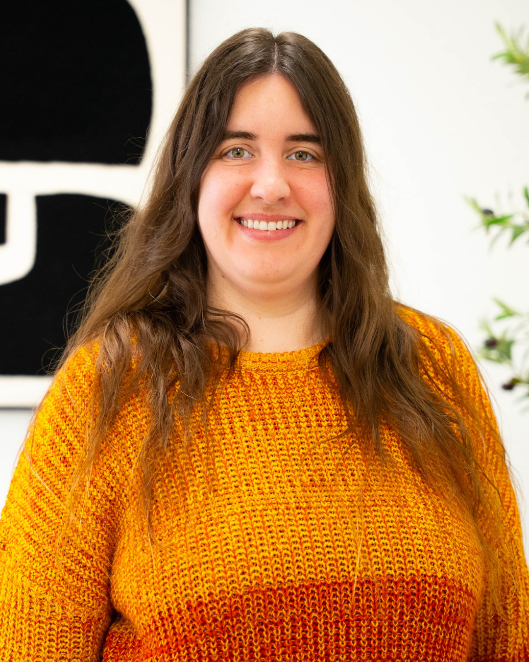 A woman with dark brown hair, dark eye brows, and light green eyes wearing an ombre orange sweater. She's standing in front of a black and white art print and a decorative olive tree.