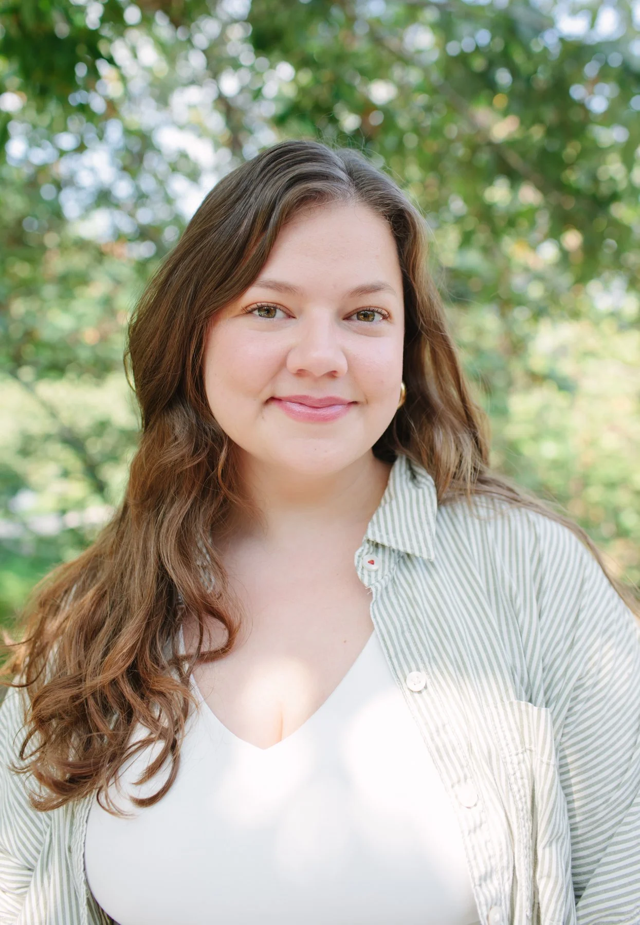 a woman with medium dark brown hair and hazel eyes stands outside in front of a tree wearing a olive green and white striped button down shirt left unbuttoned over a white v neck tank top.