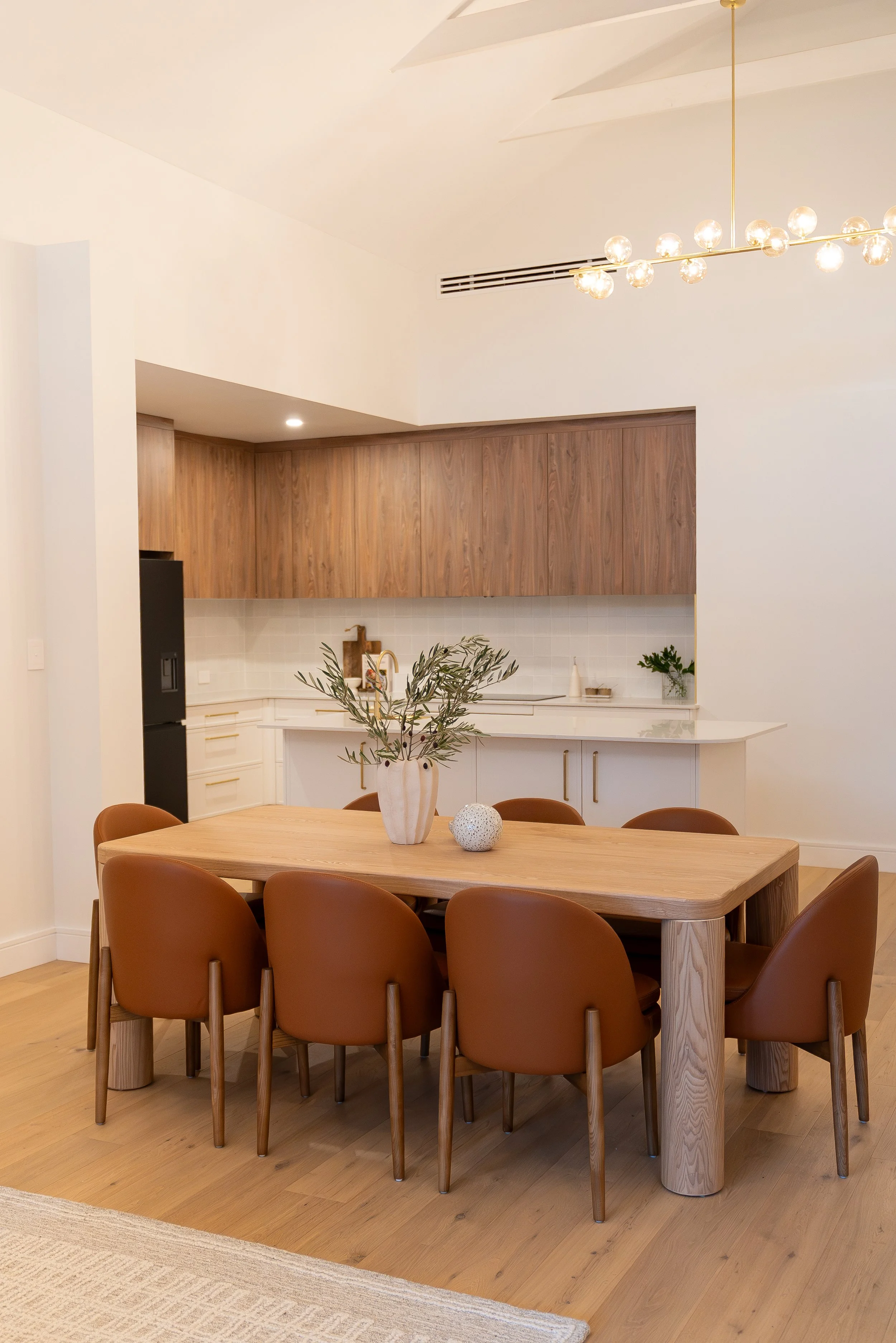 A dining room with a wooden table, brown chairs, a vase with greenery, and a modern chandelier, with a kitchen in the background.