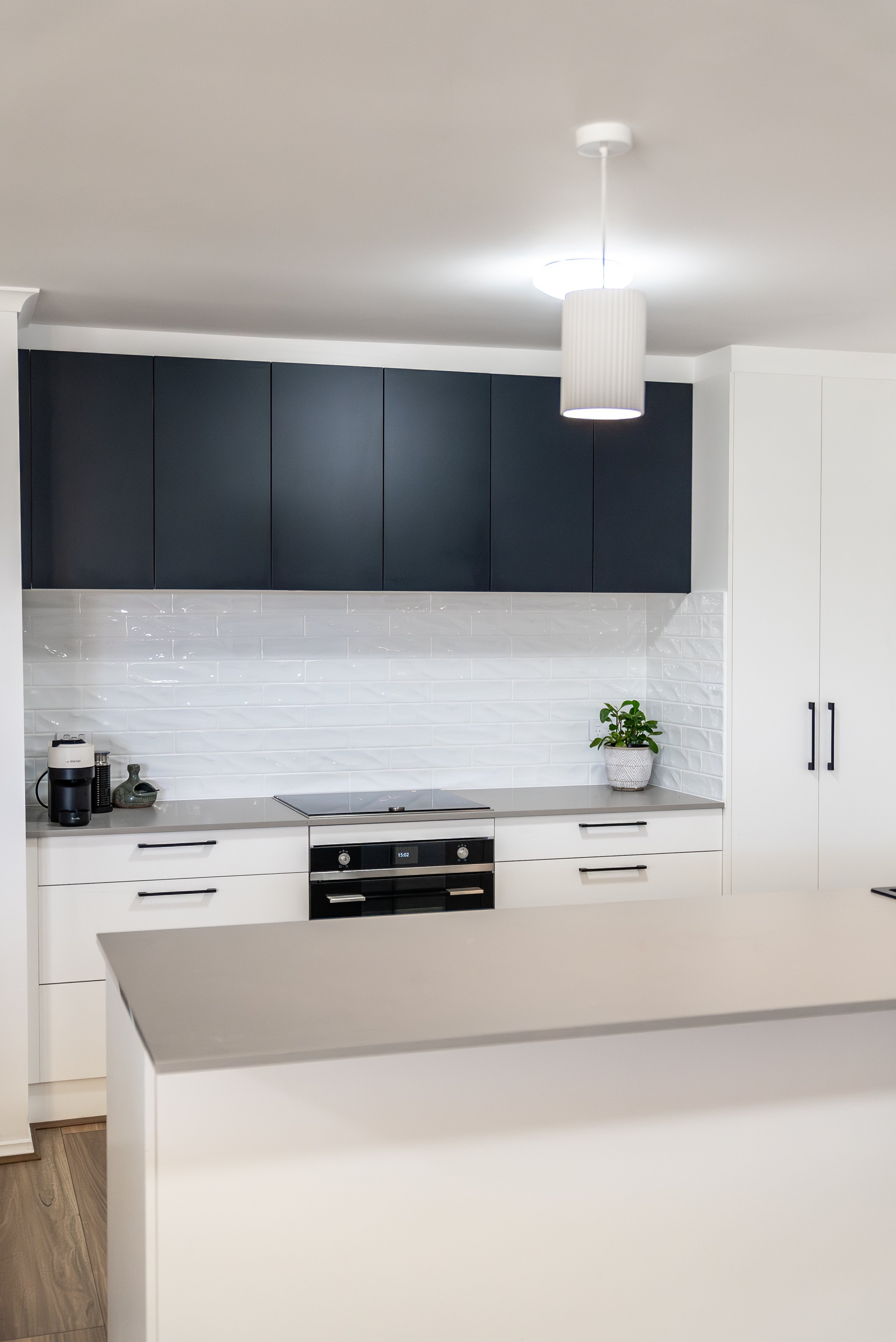 Modern kitchen with black upper cabinets, white lower cabinets, a white tile backsplash, a built-in oven, a potted plant on the counter, and a ceiling light fixture.