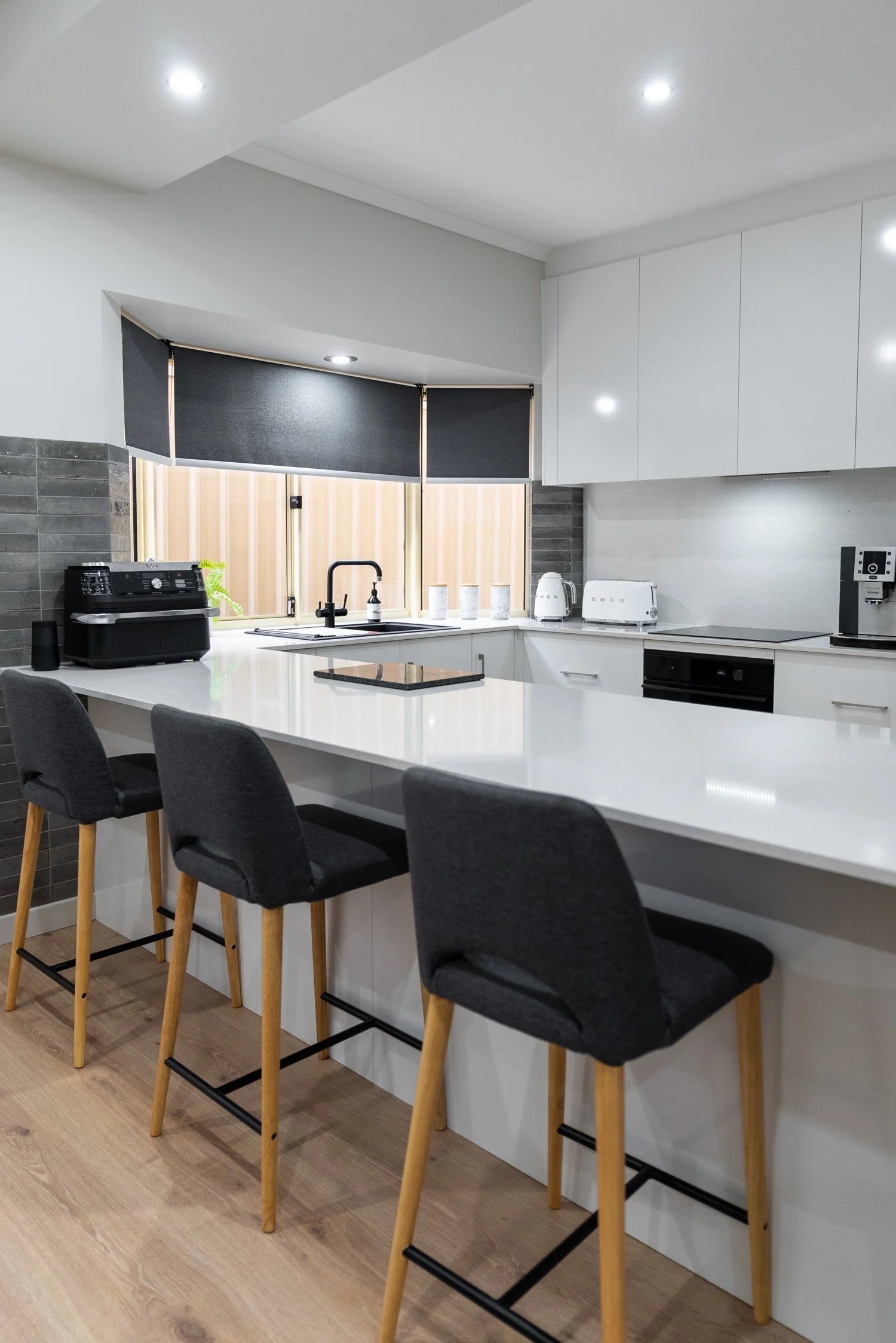 Modern kitchen with white cabinets, a large white countertop island with four black upholstered bar stools with wooden legs, black window shades, and various small appliances on the counter, including a toaster, kettle, and coffee maker.