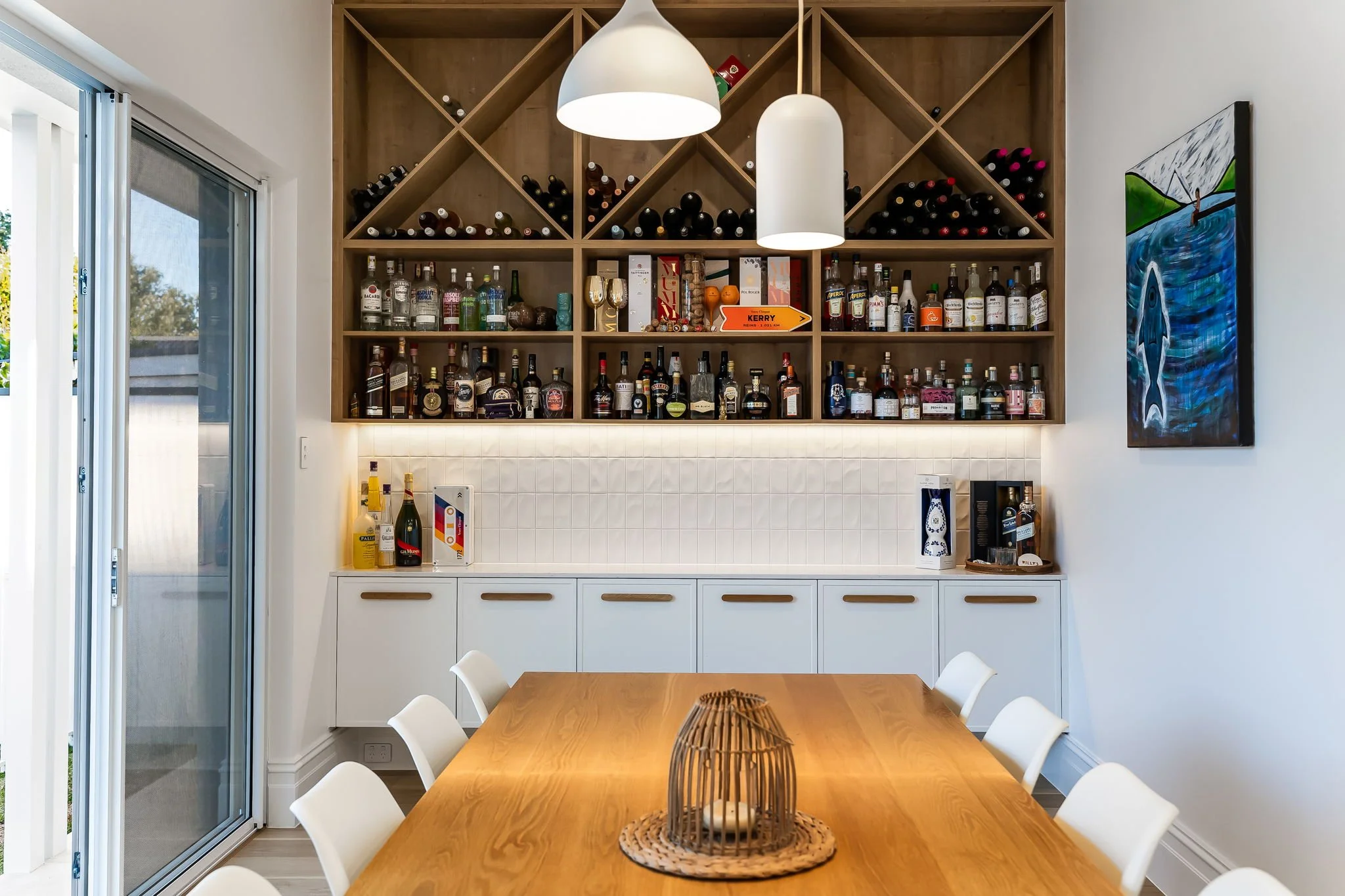 A wooden dining table with six white chairs, a decorative lantern centerpiece, a white cabinet with bottles and boxes, a wooden wine rack with bottles, liquor bottles on shelves, a painting of a fish on the wall, and a sliding glass door leading outside.