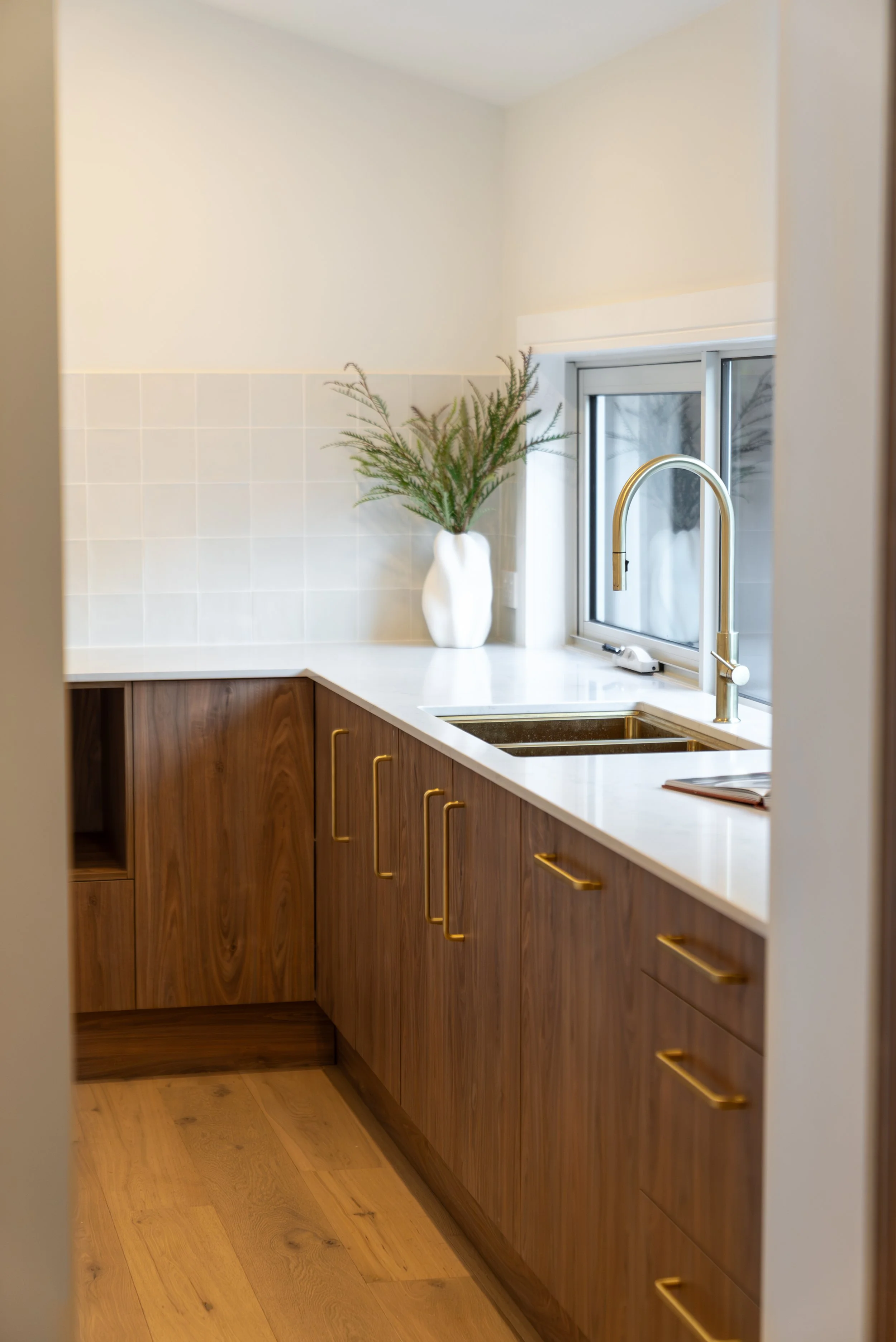 A modern kitchen corner with wooden cabinets, a white countertop, a brass faucet, a window, a white decorative vase with greenery, and a cutting board on the sink.