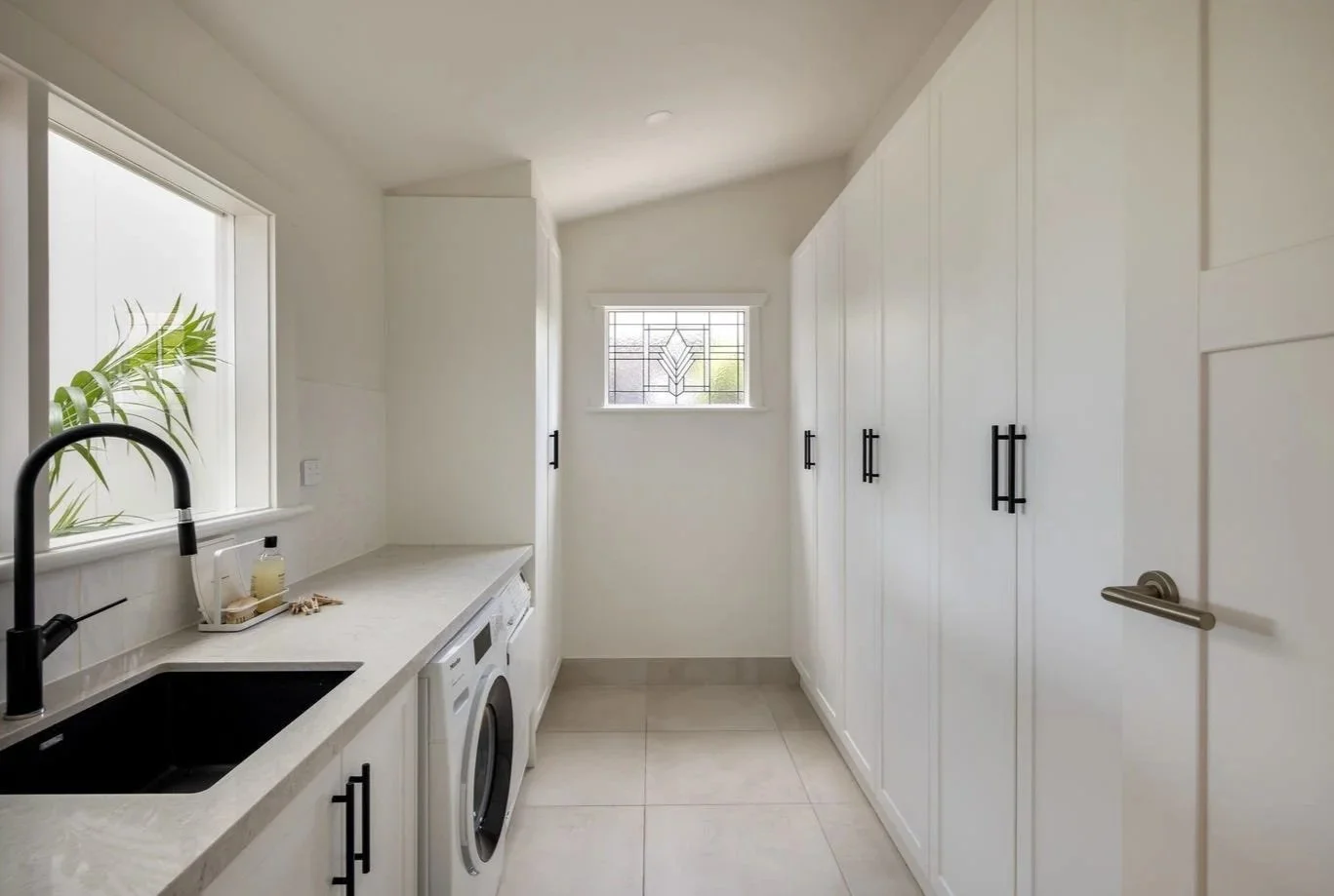 Laundry room with white cabinets, a black sink, a washing machine, two small windows, and modern black handles.
