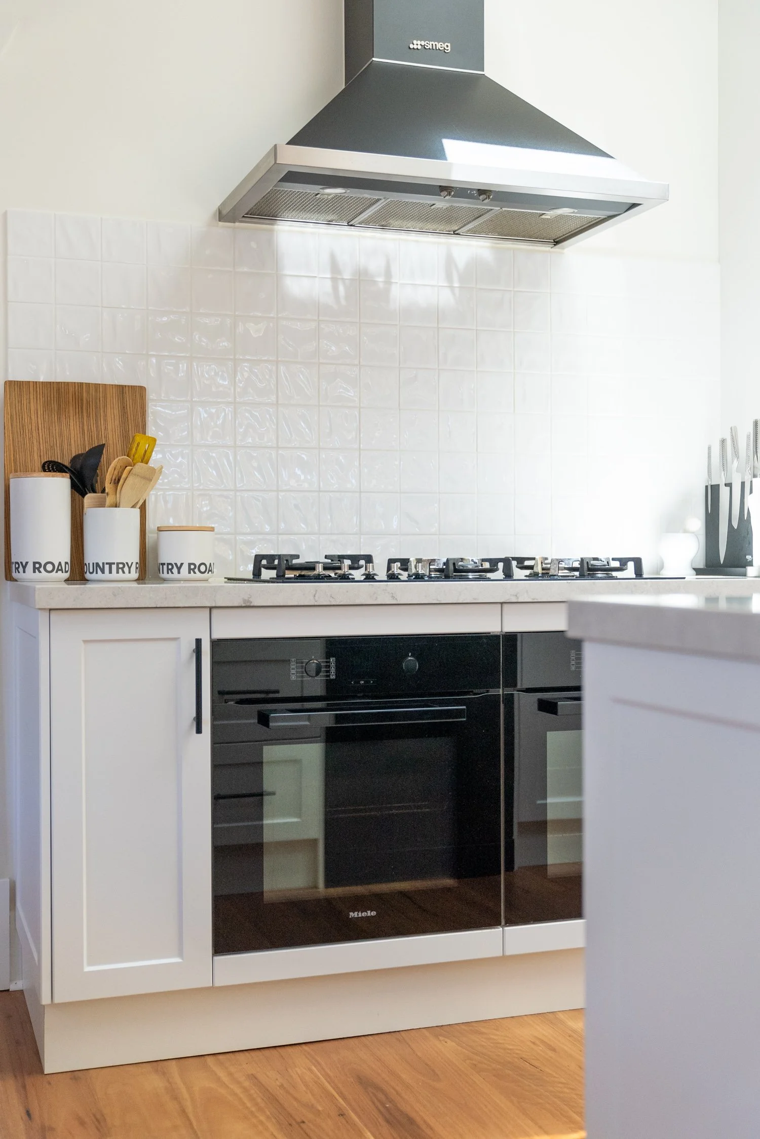 Modern kitchen with white cabinets, a black oven, a gas stove, and a stainless steel range hood. Decorative containers labeled 'Country Road' sit on the countertop.